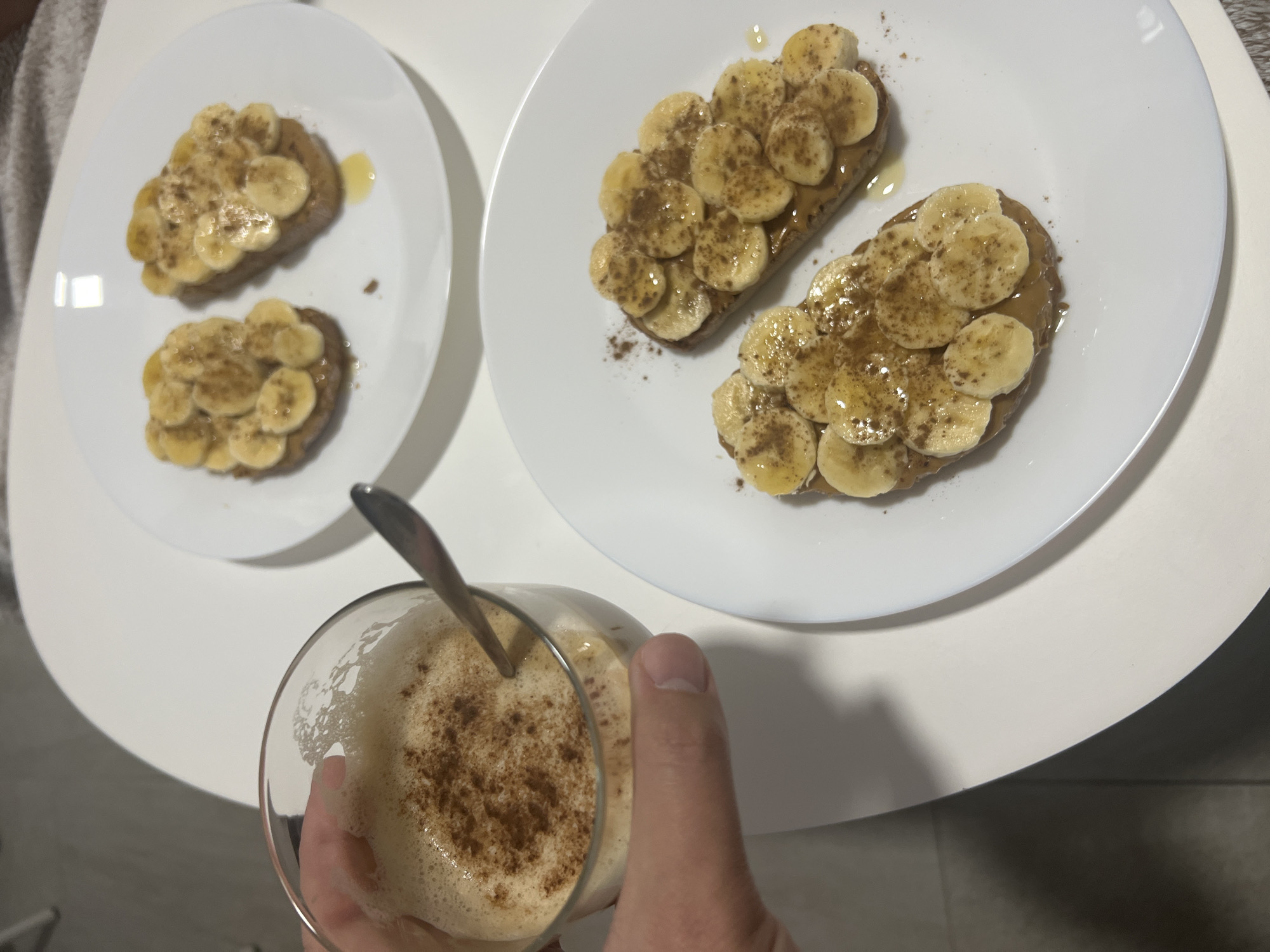 The image shows two white plates of banana toast on a white round table. Each piece of toast is covered with what appears to be peanut butter, sliced bananas, and a sprinkle of cinnamon. A glass of foamy beverage with a spoon, held by a hand, sits in the foreground. The scene takes place indoors, with a glimpse of the tiled floor and a grey garment suggesting a domestic setting. The location in Mogán, Spain is not discernible from the image.