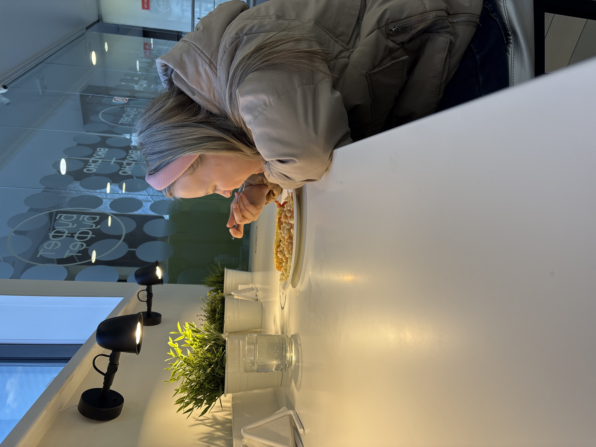 A woman is seated at a white table, eating pasta from a white plate. She is wearing a beige puffy jacket and a pink headband. Her blonde hair is visible. She is using cutlery to eat. On the table, next to her plate, is a glass of water, napkins, and several small potted plants. Behind her is a glass wall with Cyrillic text on it, which translates to "Delicious Food". Two black lamps are attached to the wall, shining light on the plants. This scene is located in Izhevsk, Russia, as evidenced by the Cyrillic text.
