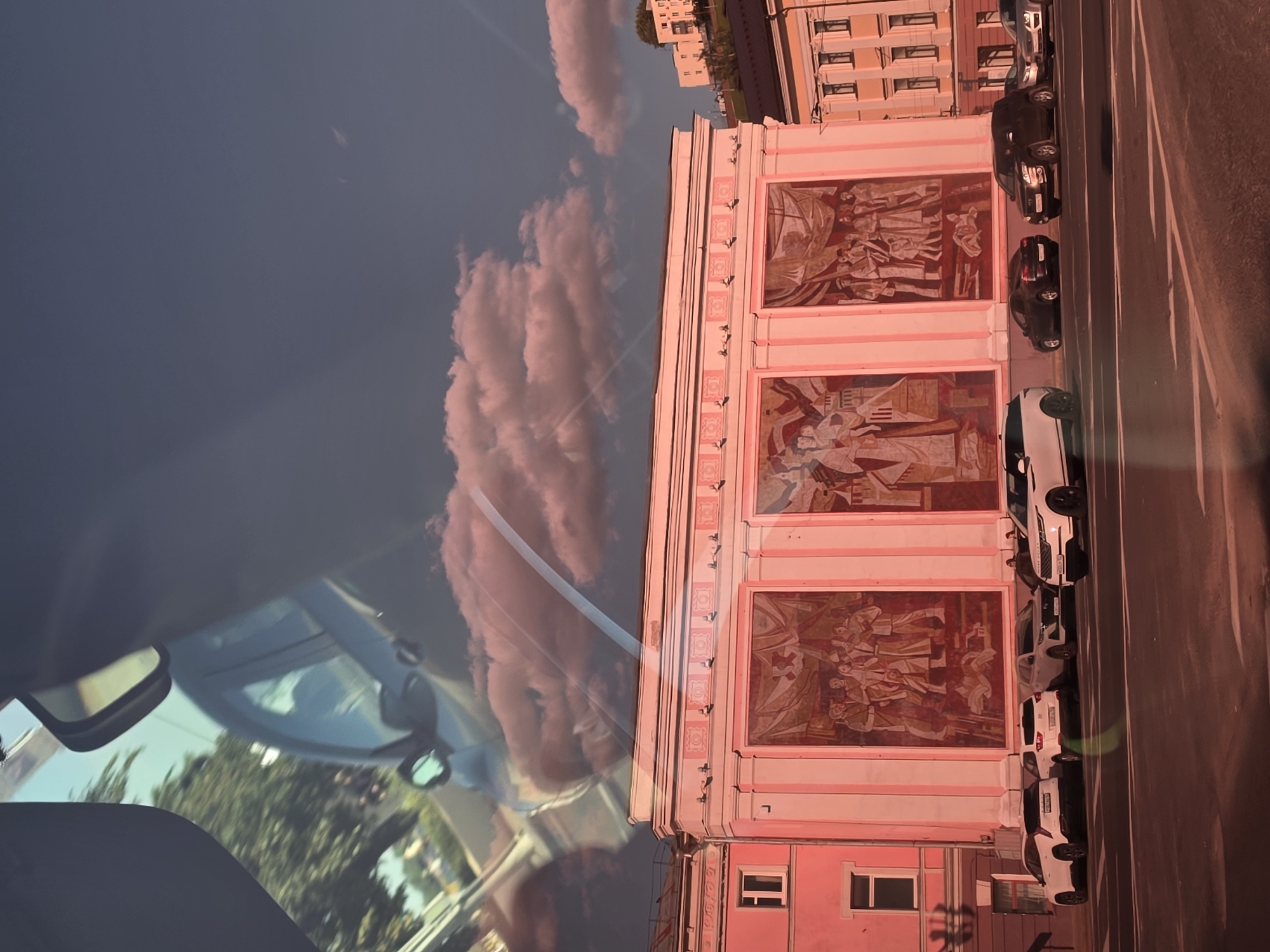 A reflection on a car windshield shows trees and a vehicle, superimposed on a view of a building with three large murals. The building is painted pink and white, and the murals depict scenes with many figures. Several white and dark-colored cars are parked on the street in front of the building. The sky above is a deep blue with some pinkish clouds. The overall scene is illuminated by the warm, reddish light of sunset, casting a reddish hue across the entire image. The location is the Nizhny Novgorod State Academic Opera and Ballet Theatre, located on Bolshaya Pokrovskaya Street in Nizhny Novgorod, Russia.