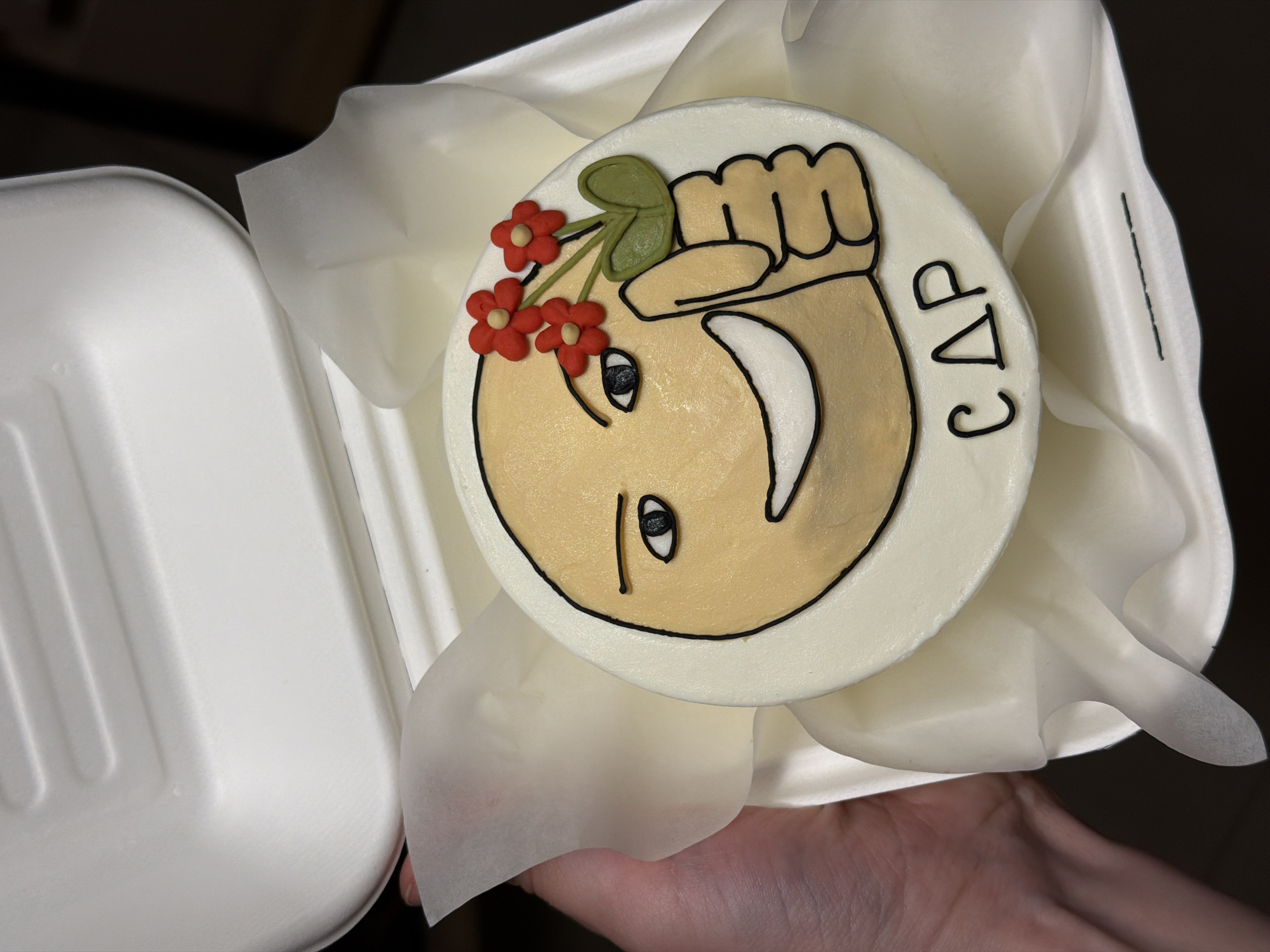 A round cake decorated with an illustration of an upside-down emoji-style face holding a bouquet of red flowers is presented in an open, white take-out container held in a hand. The cake is iced with a white base and features a smiling face with black outline, filled in with a tan color, along with red flowers, a green leaf, and the letters "C Δ P" in black. The take-out container has white tissue paper lining the edges.