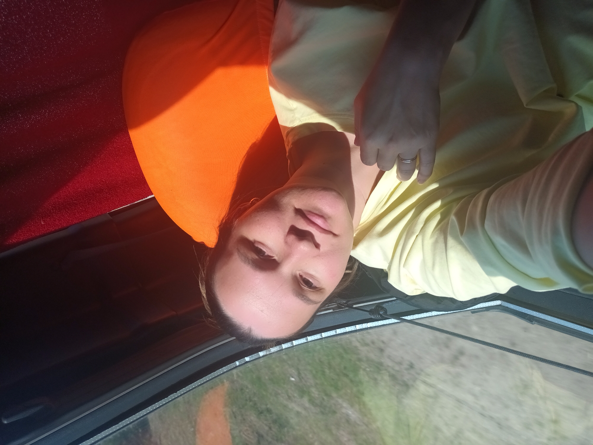 A woman is in a vehicle in Полозово, Russia. She is wearing a light yellow shirt and a ring on her left hand. She is looking at the camera. The vehicle appears to be moving, as seen through the windshield, and there is an orange pillow on the back seat. The interior of the vehicle is red and black.