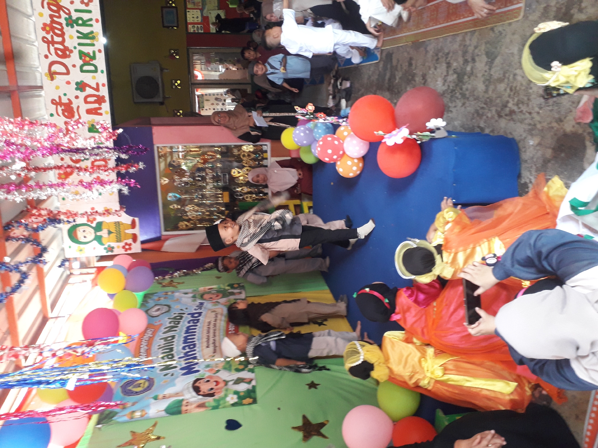 The image captures an outdoor event in Cankuang, Indonesia, likely a celebration for Mawlid Nabi Muhammad SAW. In the foreground, several children are dressed in colorful Islamic attire. On the left, a child in a yellow and orange costume with a yellow sash is seen from behind, possibly participating in a performance. Next to them, another child in a red and pink costume with a black head covering is also viewed from behind. A third child in a yellow and black costume with a gold decorative headpiece is partially visible on the right. A person in the front right, wearing a blue and white outfit, is using a smartphone to record the event. In the middle ground, several children are standing in a line. One boy in a blue shirt and grey pants, wearing a white cap, is looking to the right. Beside him, a boy in a light brown outfit and a black head covering stands with his hands clasped. Further right, a boy in a light peach-colored outfit and a black cap is smiling and looking towards the right, appearing to be a performer. Another boy in a white outfit is visible in the background, facing away from the camera. The backdrop features a green banner with cartoon depictions of children and mosques, and text related to the Mawlid celebration. Colorful balloons are scattered around the stage area. In the background, a woman in a hijab is holding a piece of paper. The environment appears to be a decorated outdoor space, possibly a schoolyard or community area, with a roof structure and various decorations, including streamers and what appears to be a display of trophies behind a glass enclosure. The ground is a mixture of concrete and dirt.