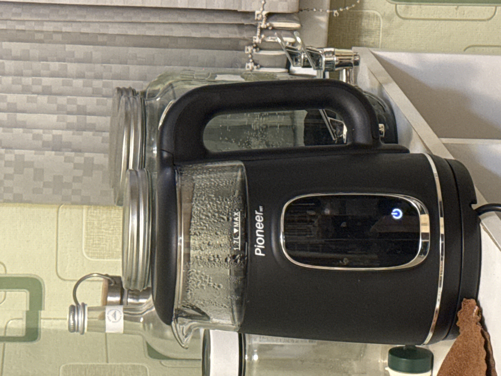 An indoor close-up shot features a black and clear electric kettle, illuminated power button on, resting on a white surface. The kettle, branded "Pioneer HT", displays "1.7L MAX" near its clear glass section, which shows condensation on the inside, suggesting recent use or impending boiling. A blue-lit power symbol button is visible on its black body, framed by a silver trim.
To the left of the kettle, a glass bottle with a cork and metal stopper is partially visible, bearing a small, indistinct white label. Behind and to the right of the kettle, a large glass jar can be seen. The background features light-colored, textured wall covering or blinds with a subtle geometric pattern. A chrome faucet or tap is visible in the upper right background, suggesting a kitchen or utility area. A brown cloth or towel is partially visible to the right of the kettle. The scene is evenly lit by indoor light.