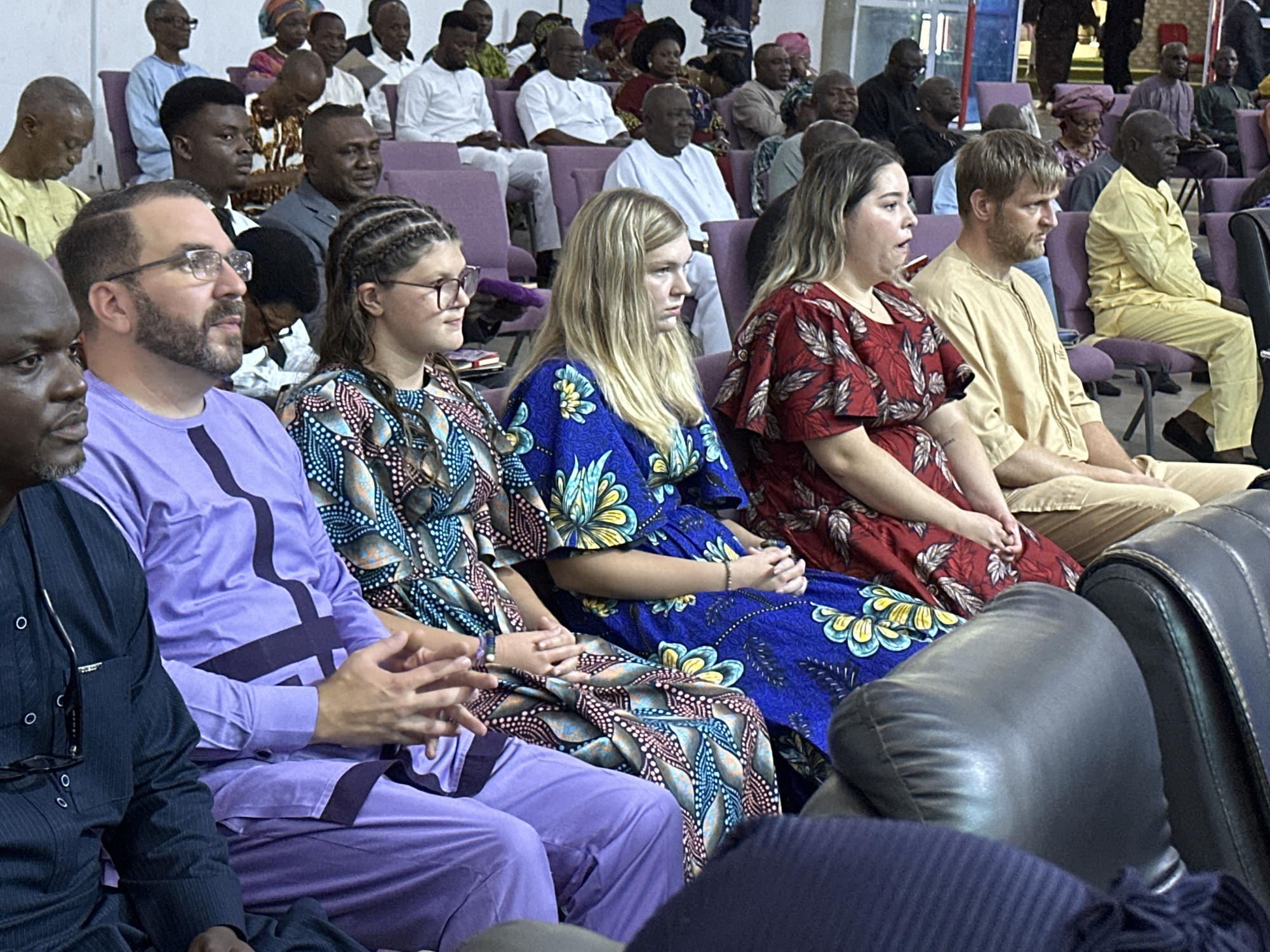 The image captures a scene inside a dimly lit indoor space, likely a church or meeting hall, with rows of purple chairs occupied by a diverse group of people. In the foreground, several individuals are seated, their attention directed forward. A man with a beard and glasses, wearing a purple tunic, is prominently featured on the left. Beside him, a young woman with braided hair, also wearing glasses, is looking to the side. Further to the right, another woman in a red floral dress is seated, and next to her, a man in a light brown tunic is looking away. The background is filled with more attendees, many of whom are wearing traditional clothing. The overall atmosphere appears to be one of attentive observation or participation in an event.