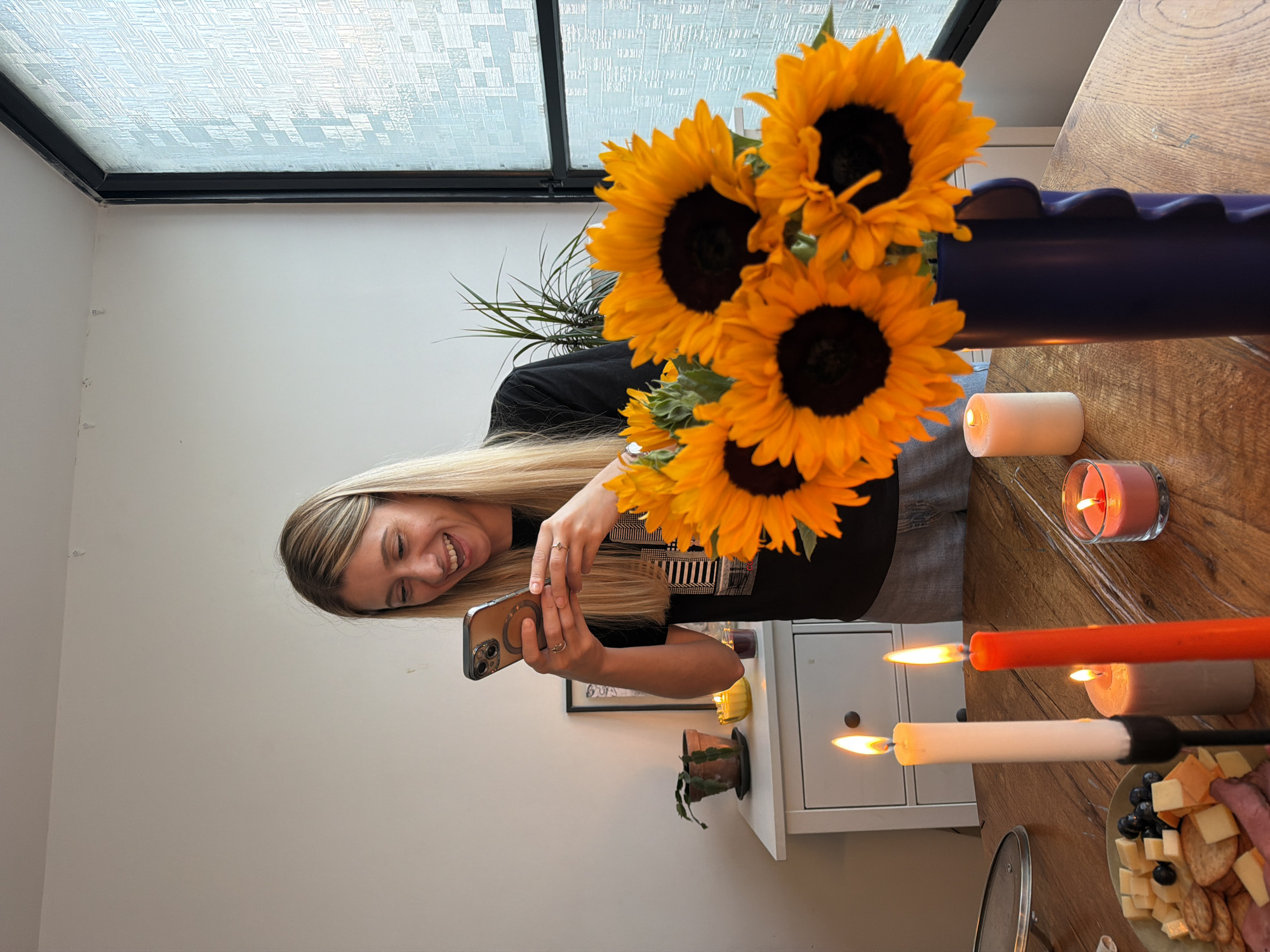 A woman with long blonde hair is smiling while taking a picture of herself with her phone. She is wearing a black t-shirt and jeans. In front of her is a bouquet of sunflowers in a dark purple vase. On the wooden table, there are three lit candles, one white, one orange, and one pink. There is also a plate of cheese, crackers, and fruit. The scene appears to be indoors, with a large window in the background that lets in natural light. The overall mood is cheerful and relaxed. The image has been rotated 90 degrees clockwise.