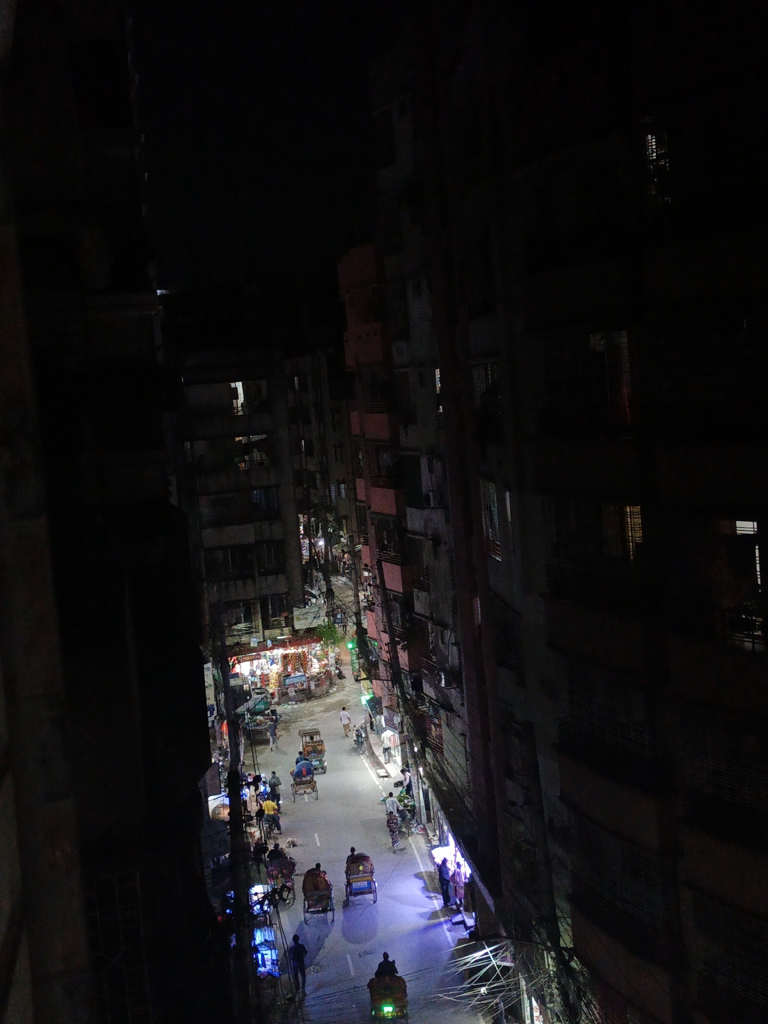 Captured from a high vantage point at night, the image reveals a narrow urban street flanked by tall, densely packed buildings in an unknown city.
The street is bathed in artificial light from storefronts and streetlights, revealing a variety of rickshaws and motorized vehicles traversing the roadway. Pedestrians are visible on the sidewalks. The buildings lining the street are multi-story structures, exhibiting an older architectural style with visible signs of wear and numerous air conditioning units protruding from the walls. Power lines and cables are strung across the street. The image shows a slice of urban life in an environment with high population density.