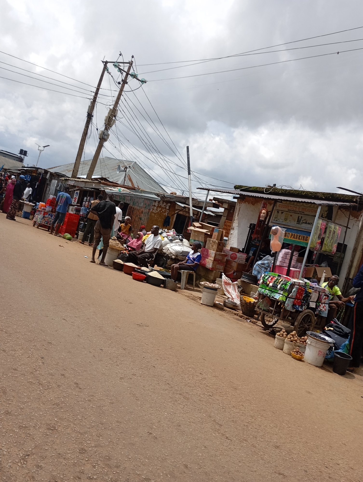 The image depicts a street market in Gura Top, Nigeria, under a cloudy sky. The scene is filled with various stalls and vendors selling goods. In the foreground, a dirt road stretches into the distance, lined with temporary structures made of corrugated metal and wood.
Several people are visible throughout the market. Some are seated, while others are standing and interacting with vendors. There are numerous goods on display, including produce such as fruits and vegetables, as well as packaged items and what appear to be clothing or textiles on a cart. Several buckets and containers are also present, holding various commodities. Power lines with multiple cables crisscross the sky above the market, supported by tall wooden poles, one of which is visibly leaning. The overall impression is one of a bustling and active local marketplace.