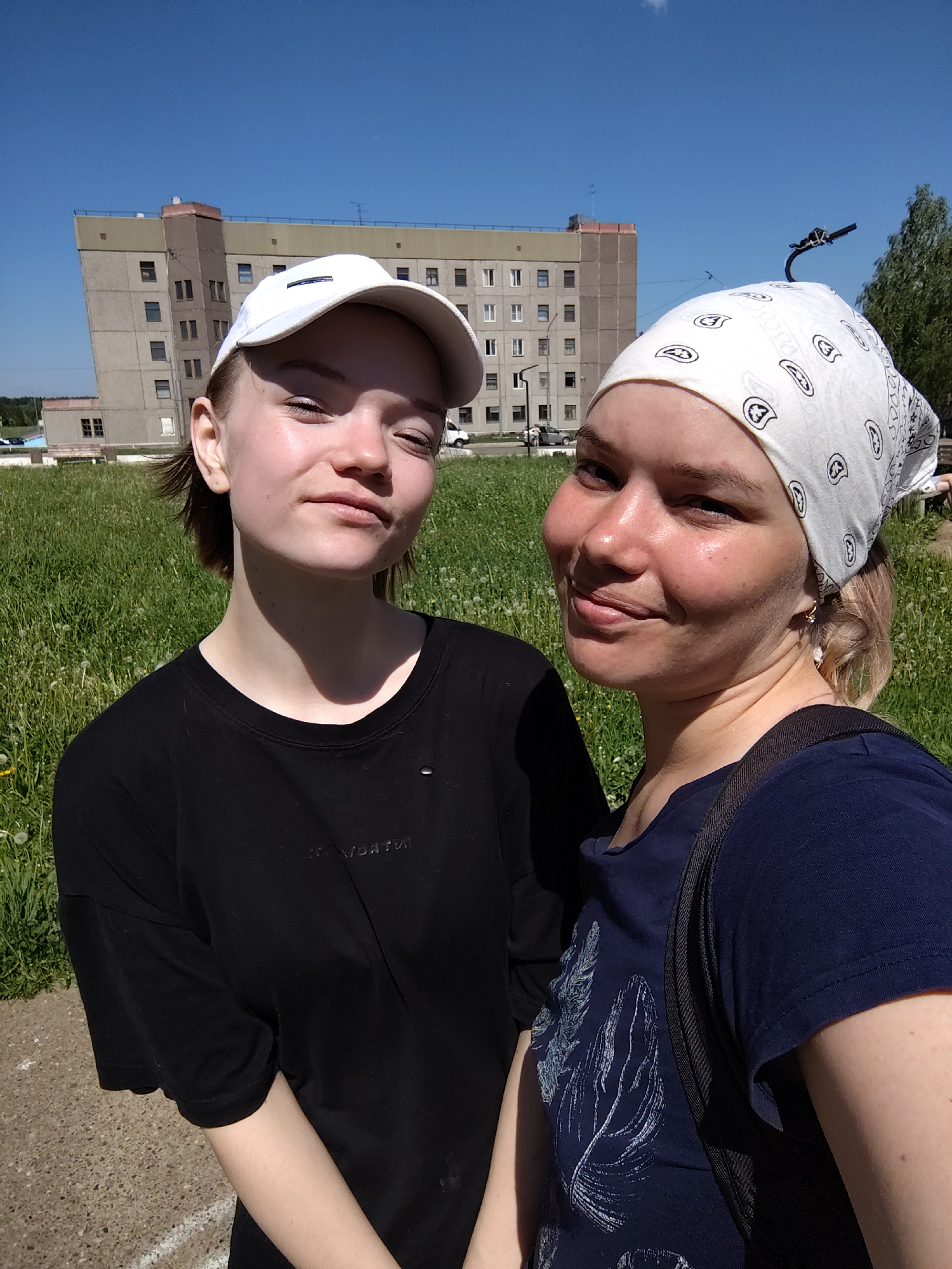 The image is a medium shot of two young women posing outdoors. The woman on the left wears a white baseball cap, a black T-shirt, and has short brown hair. The woman on the right wears a white bandana with a black paisley print. She has blonde hair, and a blue T-shirt. A black backpack strap is visible on her shoulder.
The background features a grassy field with dandelions. Behind the field, there is a large, drab building with several stories and many windows. In the distance, a blue sky is seen above the building and trees.
The location is Марковский, Russia, as the image comes from that city.