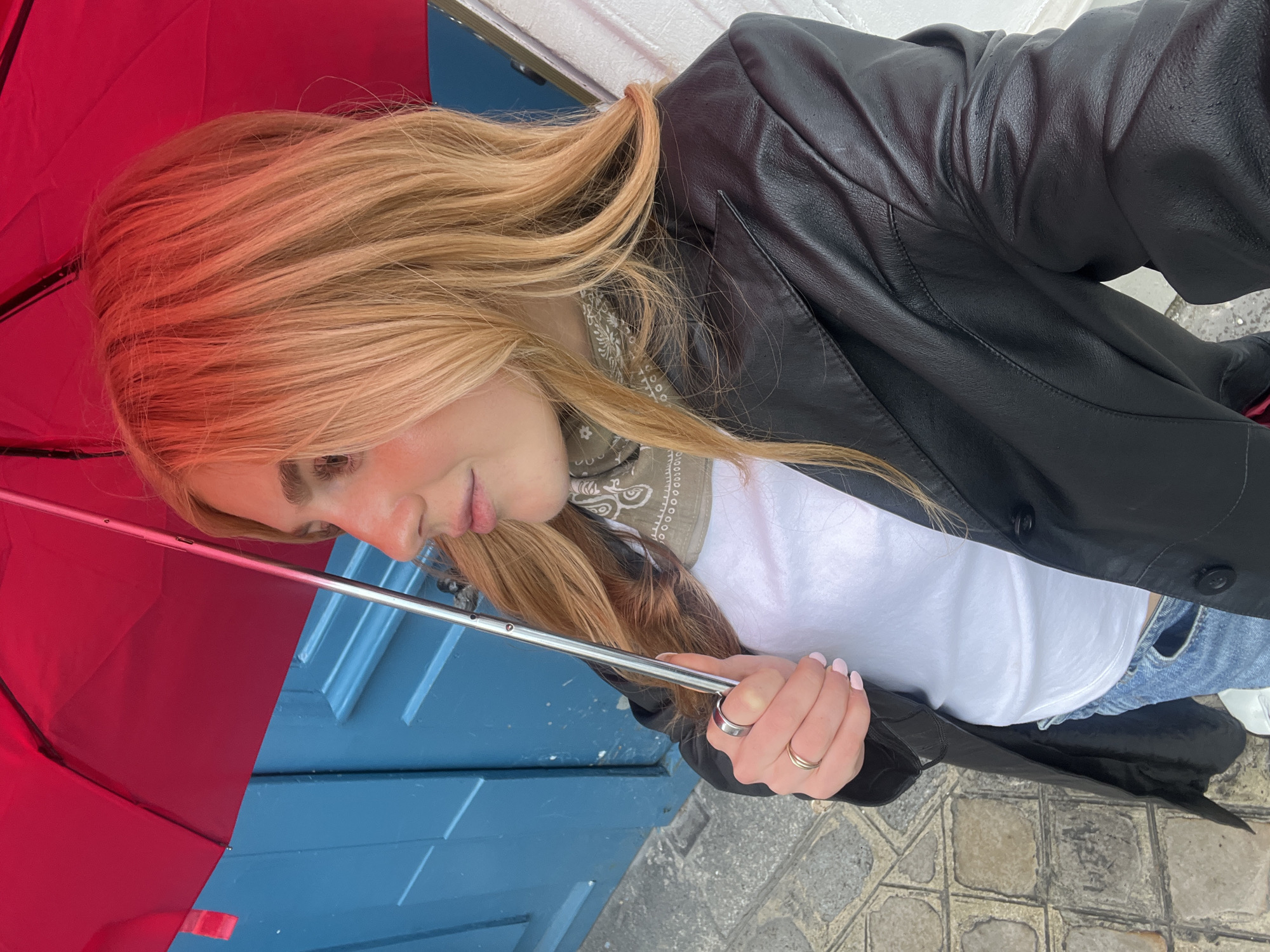 A person is holding a red umbrella in front of a blue door. They are wearing a black leather jacket, a white t-shirt, a bandana around their neck, and light-wash blue jeans. The person is standing on a cobblestone street. The door is part of a building with white trim. The location is likely Paris, France, given the architecture and cobblestone street.