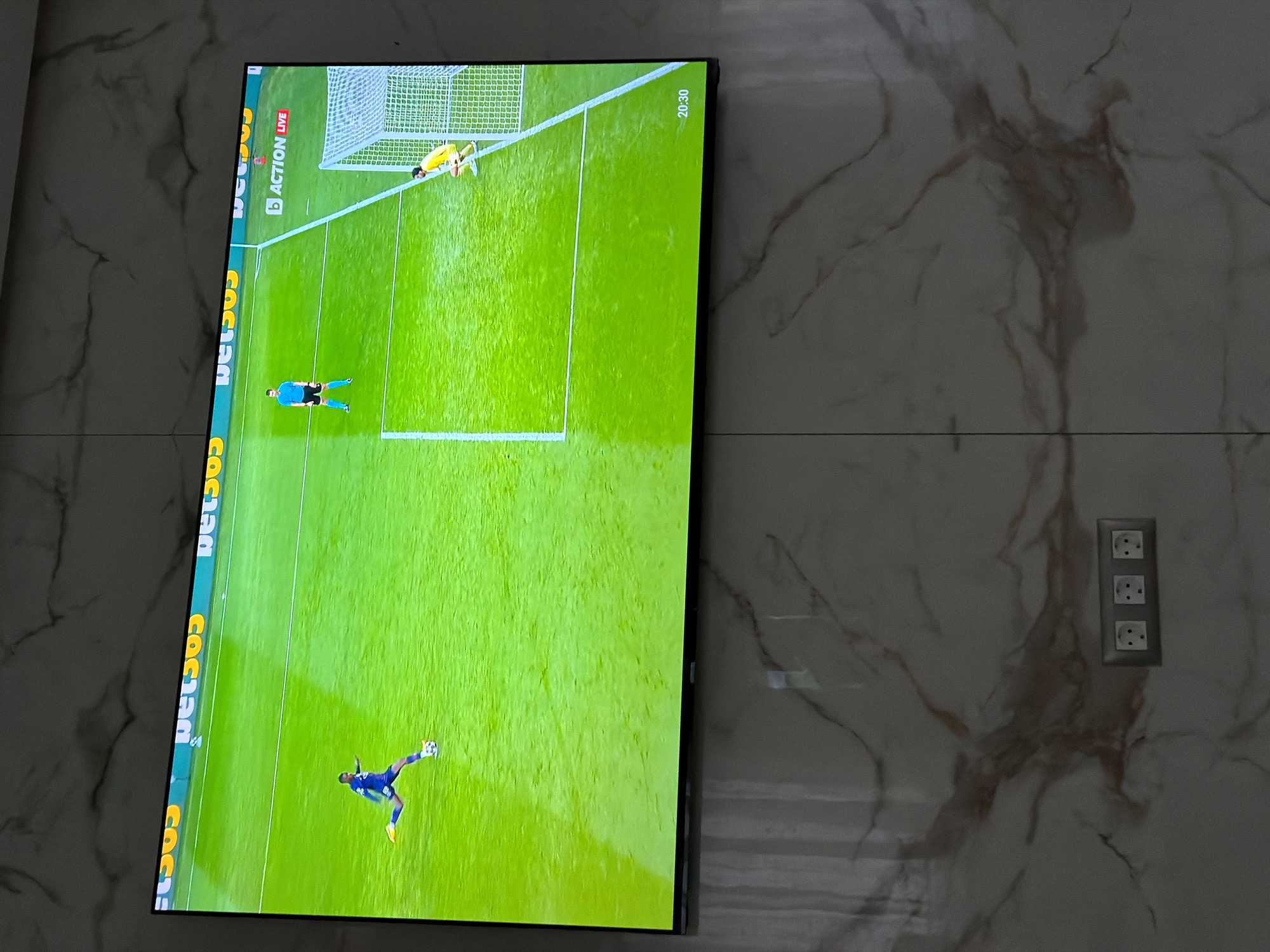 A television displays a football match on a green pitch, with players visible near a goal. The screen shows sports branding along its left edge and a time stamp "20:30" in the top right. The television is mounted on a marble-patterned wall next to a vertical arrangement of three electrical outlets. The location and its relation to Asenovgrad, Bulgaria, is not determinable from the image.