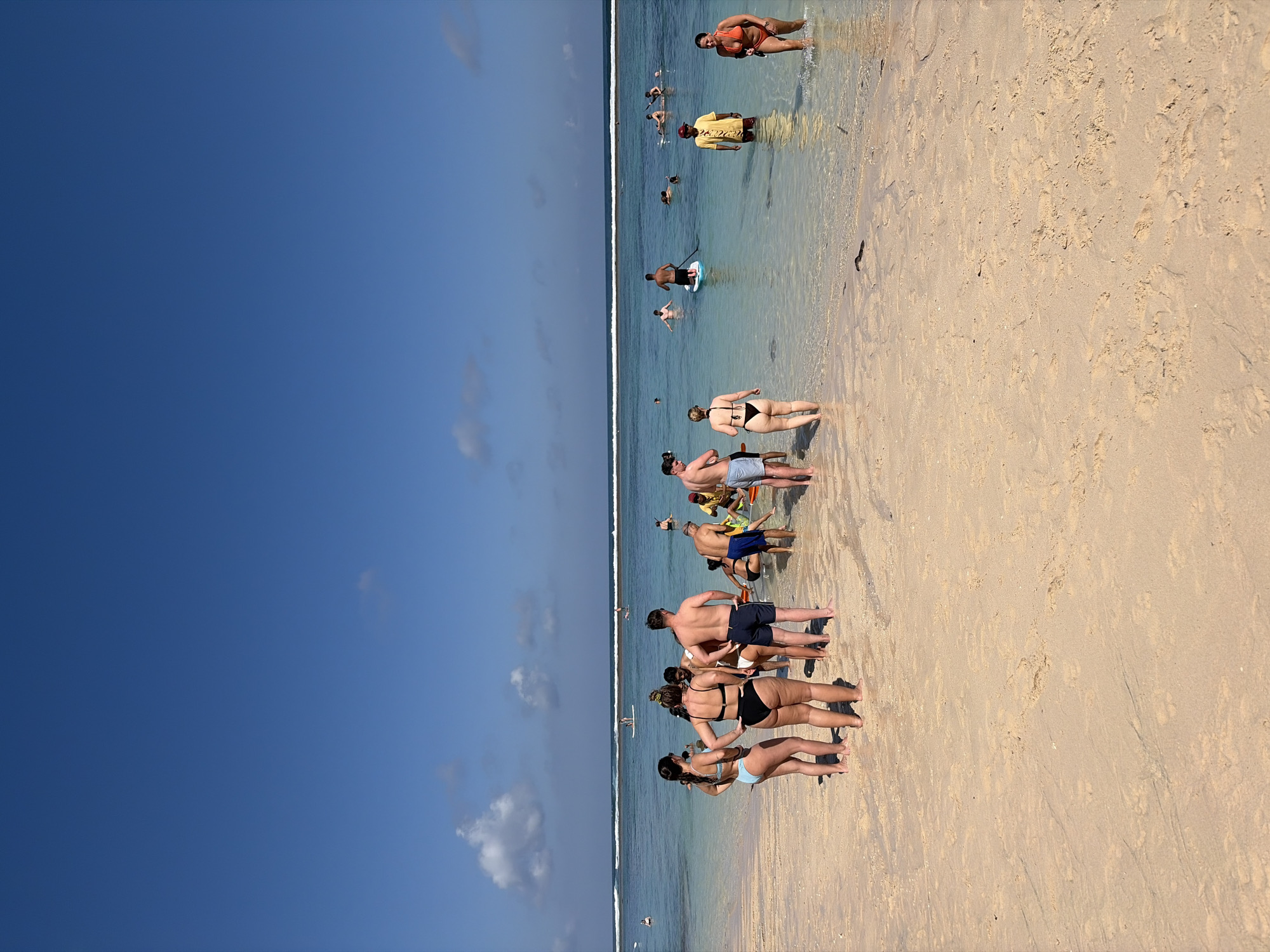 On a bright, sunny day, a group of people are enjoying a beach and shallow ocean in Kuta Selatan, Indonesia. The sand is a light tan, with gentle waves lapping at the shore. In the foreground, a group of tourists, dressed in swimwear, stand on the wet sand. Some are facing the ocean, while others are talking amongst themselves.
In the water, several people are engaged in various activities. One person is on a paddleboard, while others are swimming or wading. In the distance, more individuals are visible, with the turquoise water stretching out to meet the horizon under a clear blue sky. The overall scene is one of leisure and recreation on a tropical beach.