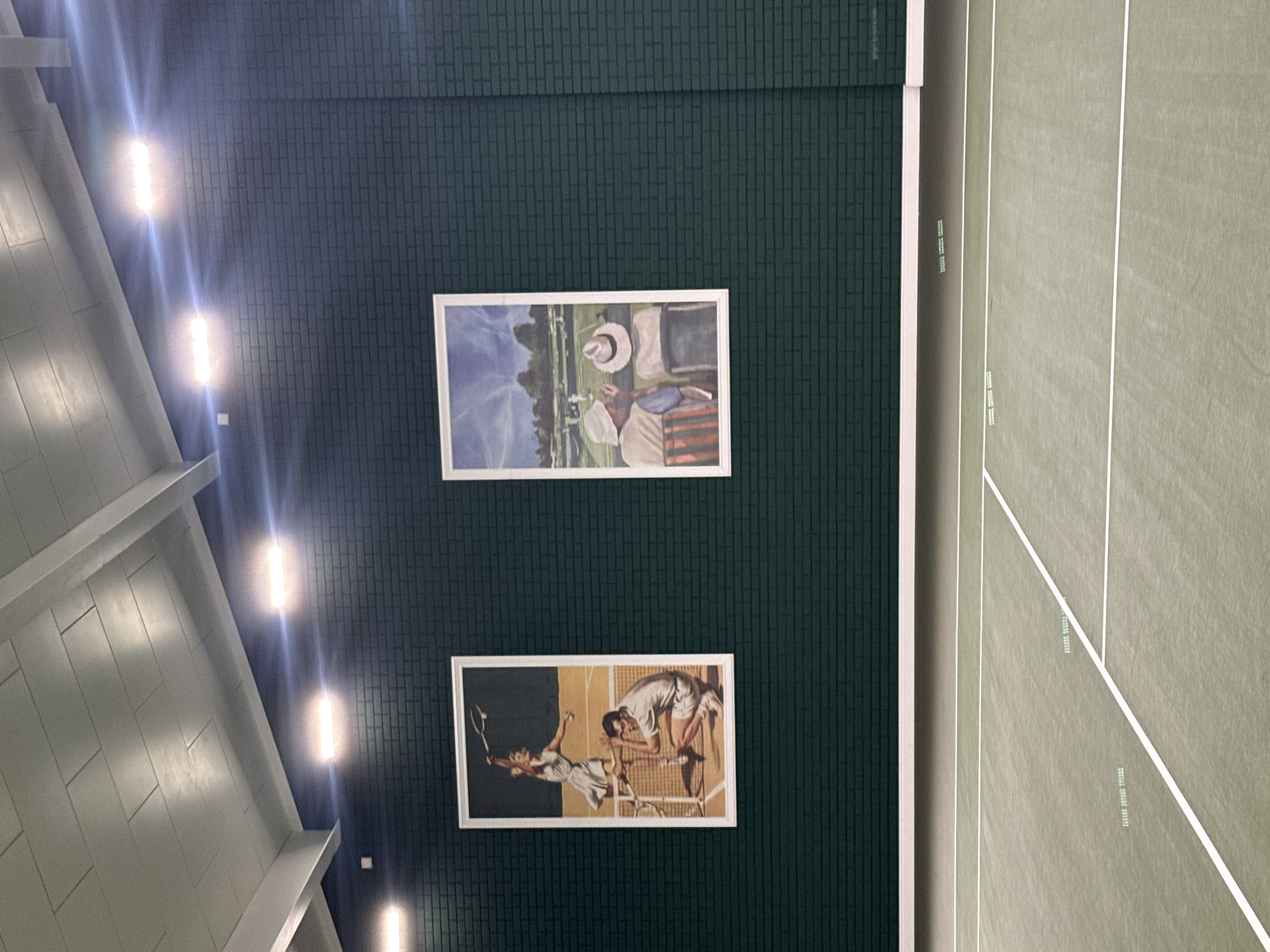 The image shows an indoor tennis court with a dark green brick wall on the left and a green court on the right. Two framed paintings are displayed on the wall. The top painting depicts two people in hats sitting on chairs facing a tennis match in a grassy field with a blue sky. The bottom painting shows a man and a woman playing tennis. The woman is in mid-air, reaching for a shot, while the man is bent over, seemingly watching the play.
The scene is indoors, indicated by the ceiling lights and the absence of natural daylight. The court appears to be well-maintained, with clean white lines marking its boundaries. The overall impression is one of a recreational facility, possibly a private club or a sports complex in Zhukovka, Russia, given the provided location context. There are no explicit indicators of time of day or weather conditions. The emotion conveyed by the paintings is one of leisure and sporting engagement. Some faint text, likely related to court specifications or branding, is visible on the court surface, but it is too blurry to read.