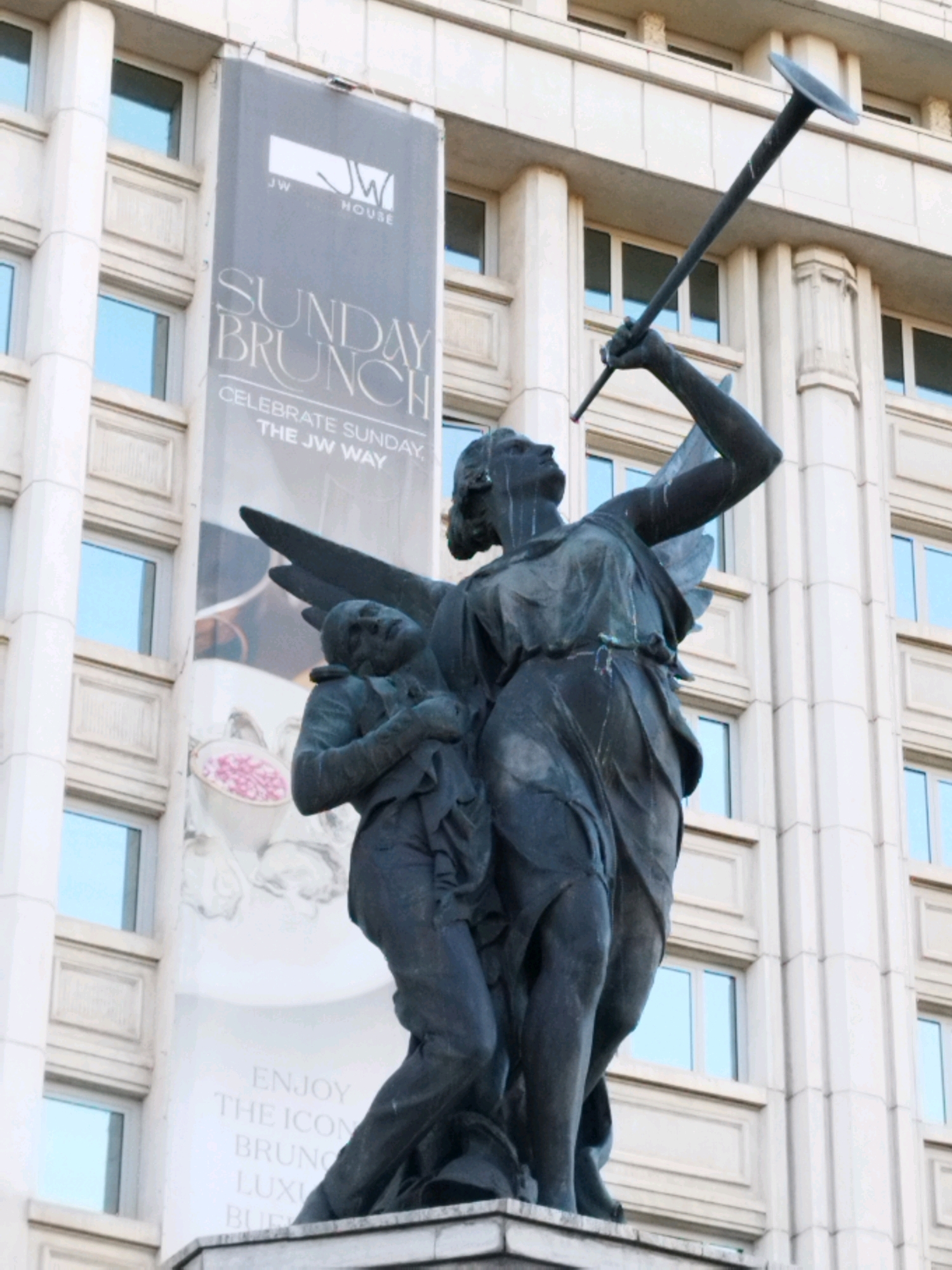 The image shows a bronze sculpture of an angel holding a long trumpet in front of a building in Bucharest, Romania. The building has a banner that says "SUNDAY BRUNCH CELEBRATE SUNDAY THE JW WAY". The sculpture depicts an angel with wings, holding a trumpet to its lips. Another figure is leaning on the angel. The angel appears to be blowing the trumpet. The building is light-colored and has many windows. The sky is not visible. The image is taken from a low angle, looking up at the sculpture and the building.