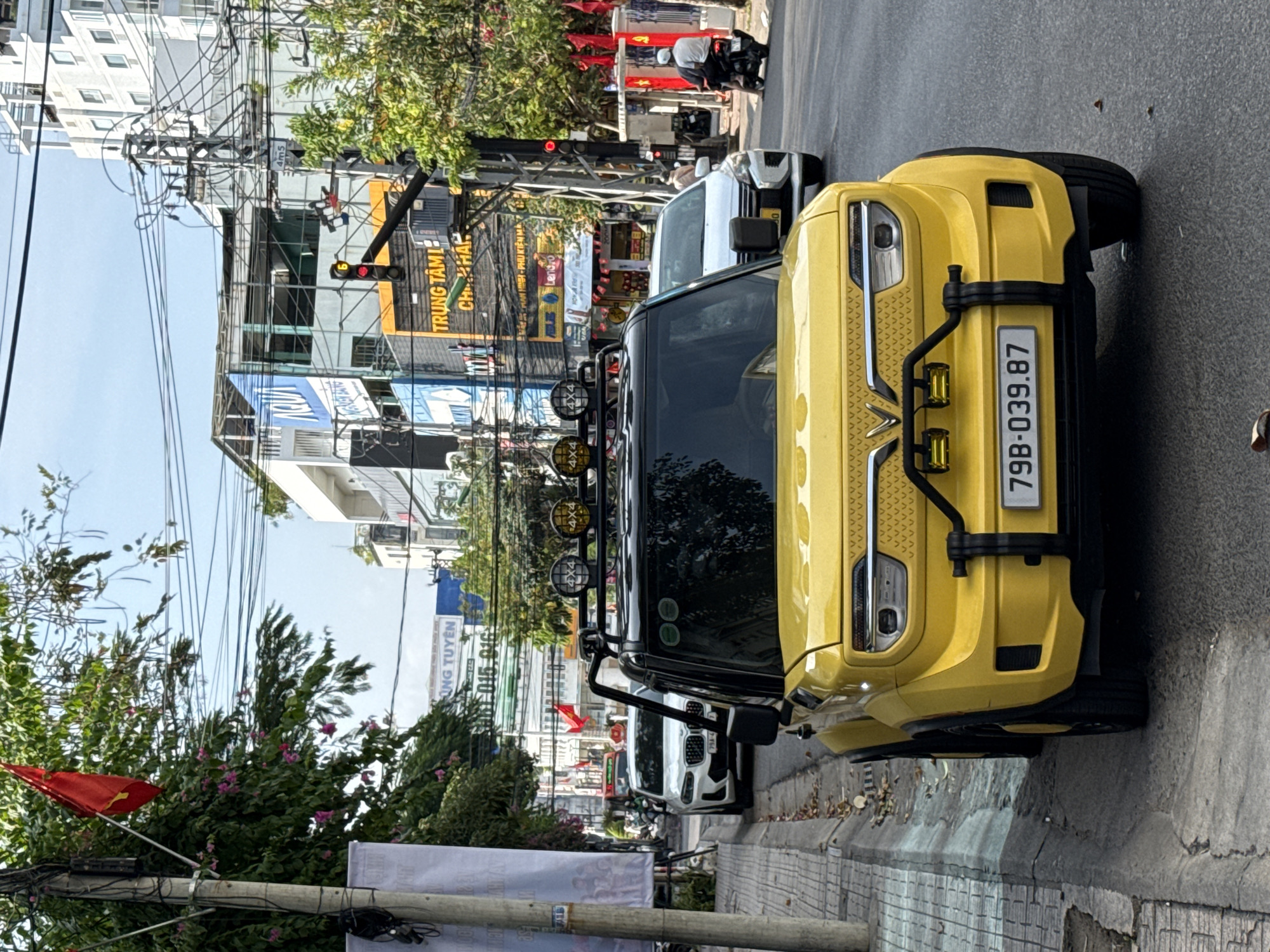 The image captures a street scene in Nha Trang, Vietnam, presenting a bright yellow car parked on the sidewalk. The car has a distinct roof rack with four auxiliary lights and a license plate reading "79B-039.87". The vehicle is parked near a flag and electrical pole. Behind the yellow car is a white vehicle. In the background are multiple businesses and restaurants with various signs and Vietnamese flags. Overhead power lines are intertwined across the sky. A traffic light is visible, showing a red signal. A person is on a scooter in the middle of the road.