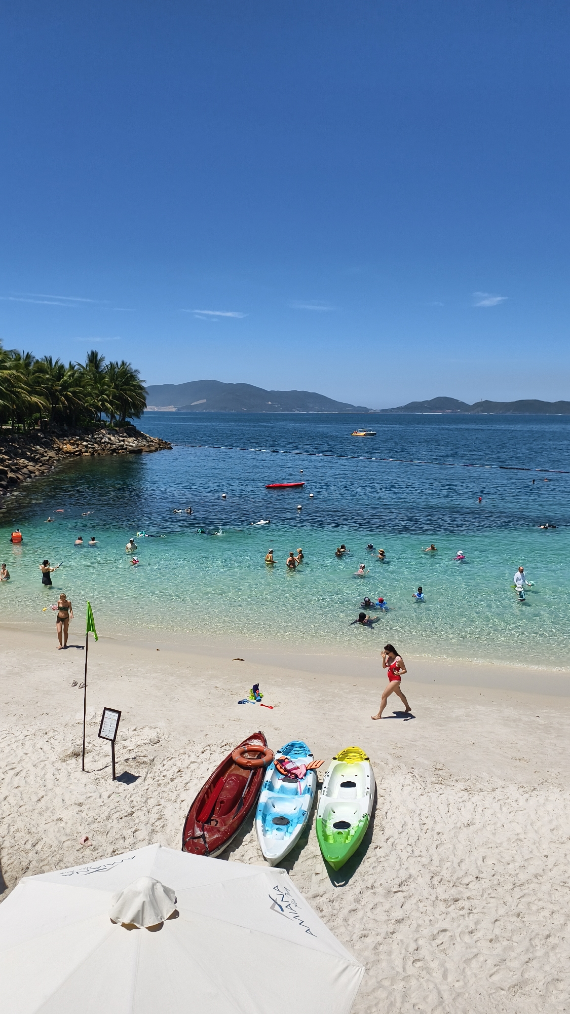 A bright, sunny day at a beach resort in Nha Trang, Vietnam. The image captures a picturesque bay with crystal-clear turquoise water, a sandy beach lined with palm trees, and lush green mountains in the background. Several people are enjoying the water, swimming, and kayaking. Three kayaks are neatly arranged on the sand, ready for use. A white beach umbrella stands open, providing shade. The overall atmosphere is one of leisure and relaxation in a tropical paradise.