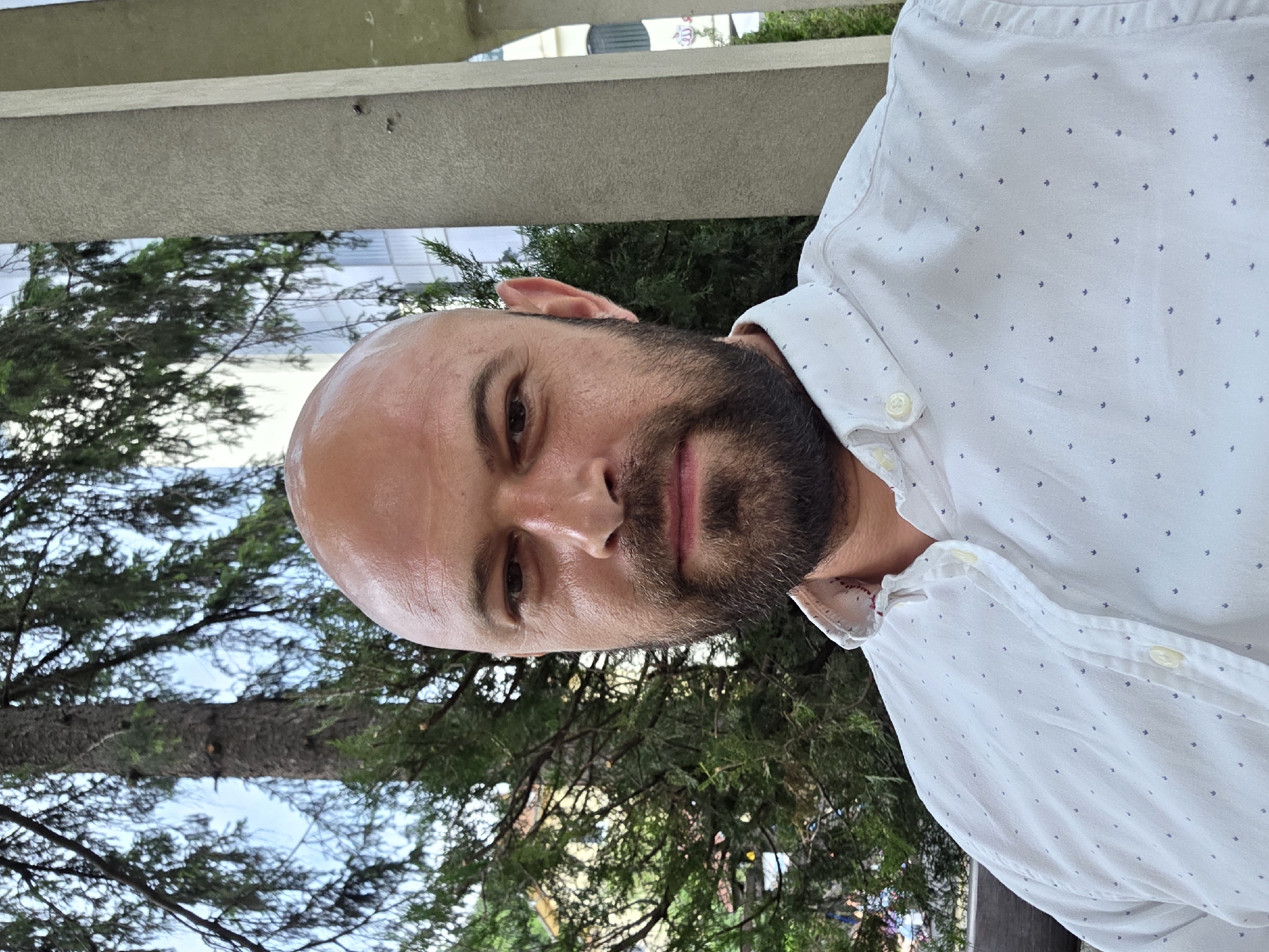 A bald, middle-aged Caucasian man with a beard is captured in a close-up, outdoor selfie. He is wearing a white button-down shirt with a subtle dark pattern. The man is positioned centrally in the frame, with his head and upper torso visible.
The background consists of greenery, likely from trees and bushes, and a blurred building. To the right of the man, a light-colored, textured pillar is visible. The setting appears to be urban, with natural elements intermingling with architectural structures. Based on the provided location information, this scene is situated in Gabrovo, Bulgaria. The lighting suggests it is daytime, possibly overcast. There are no apparent interactions or actions taking place.