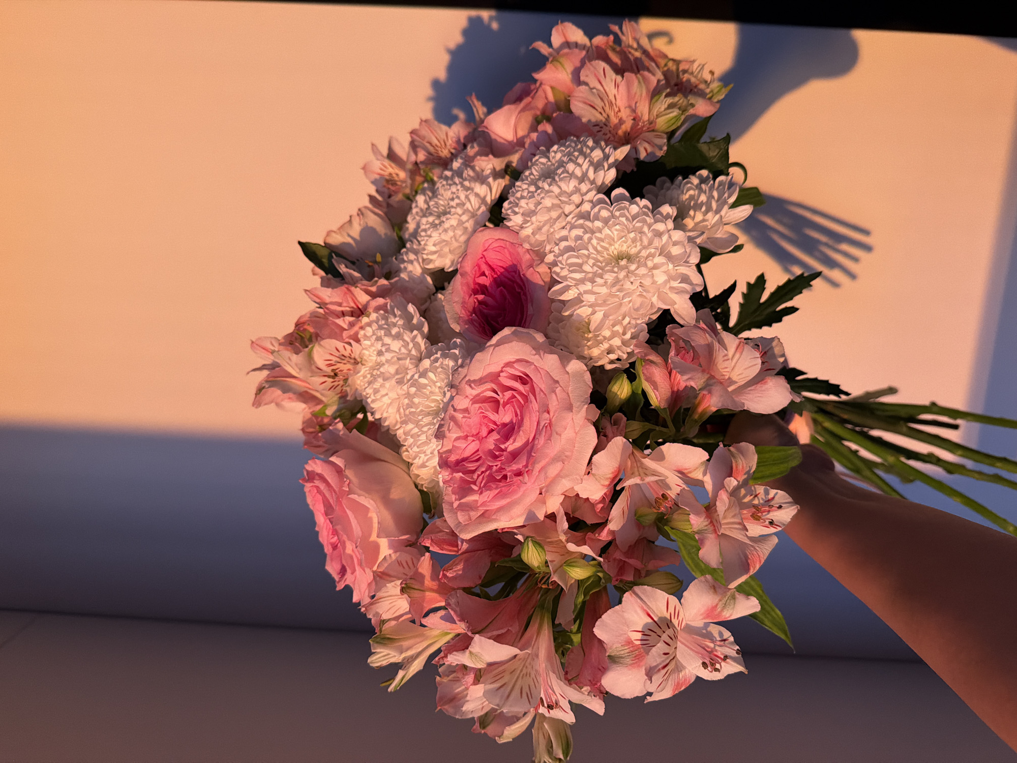 The image depicts a bouquet of pink roses, pink alstroemeria, and white chrysanthemums being held by a person's arm. The light suggests late afternoon or early evening. The setting appears to be indoors, with the background showing an ombre-like gradient going from light peach to dark blue. There is no information to confirm the image's location.