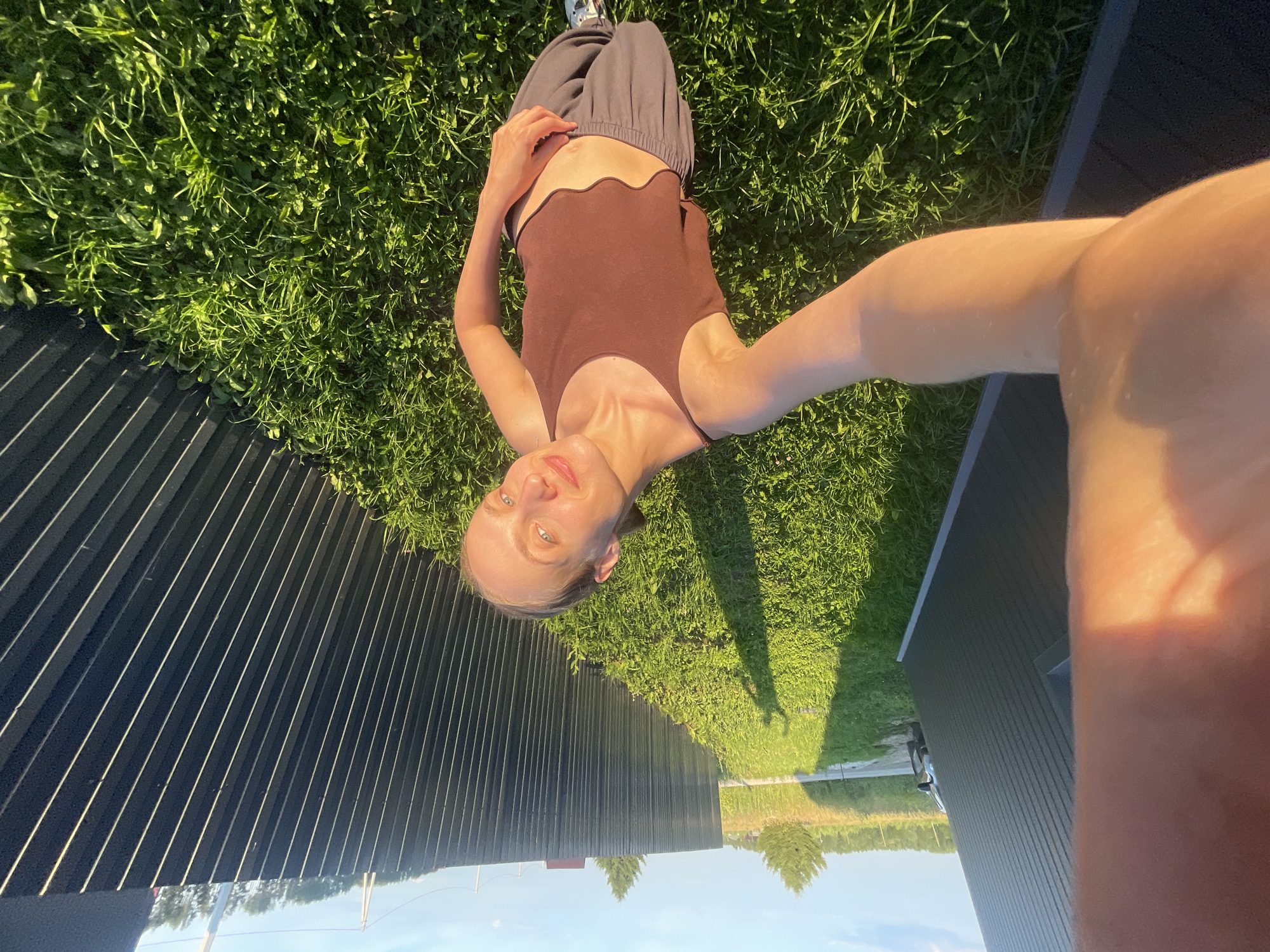In a sunlit, grassy yard, a young woman is lying on her side, taking a selfie. She has blonde hair pulled back and is wearing a brown tank top and grey sweatpants. The background features a dark corrugated metal fence, green grass, and a bright blue sky with scattered clouds. The overall setting appears to be suburban or rural. There are no discernible urban features of the city of Подшивалово, Russia, present in the image.