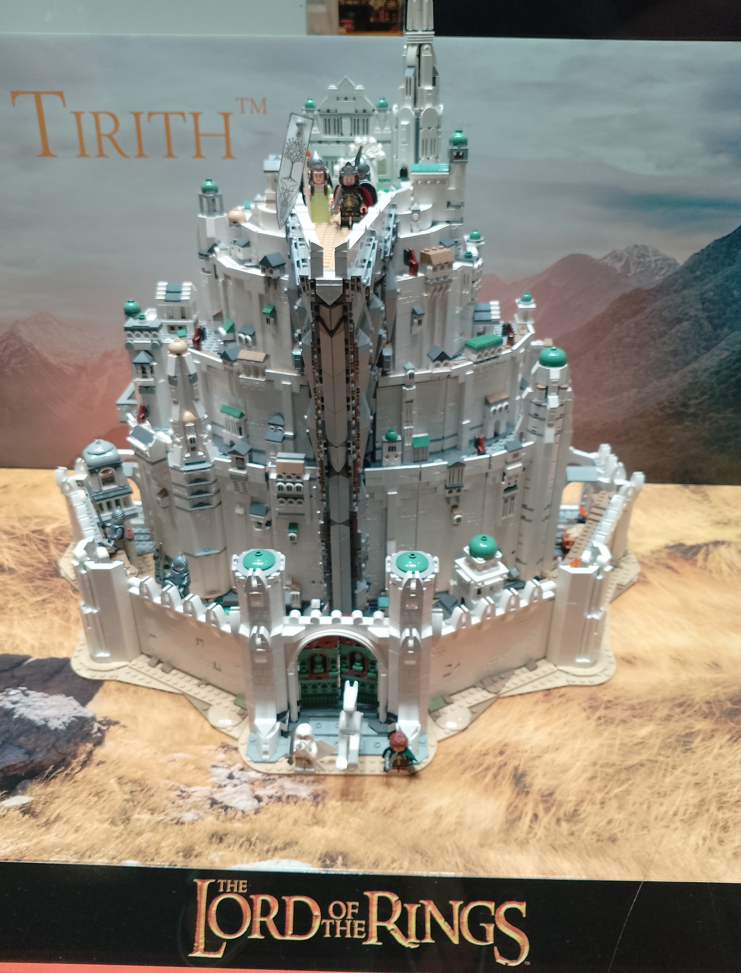 A large, white, multi-tiered Lego model of Minas Tirith from The Lord of the Rings stands on a table. The model is intricate, featuring numerous towers, arches, and small figures of people and horses. The backdrop behind the model is a scenic landscape of mountains and a hazy sky. In the foreground, the bottom edge of the image shows a black band with the logo "THE LORD OF THE RINGS" in gold lettering.