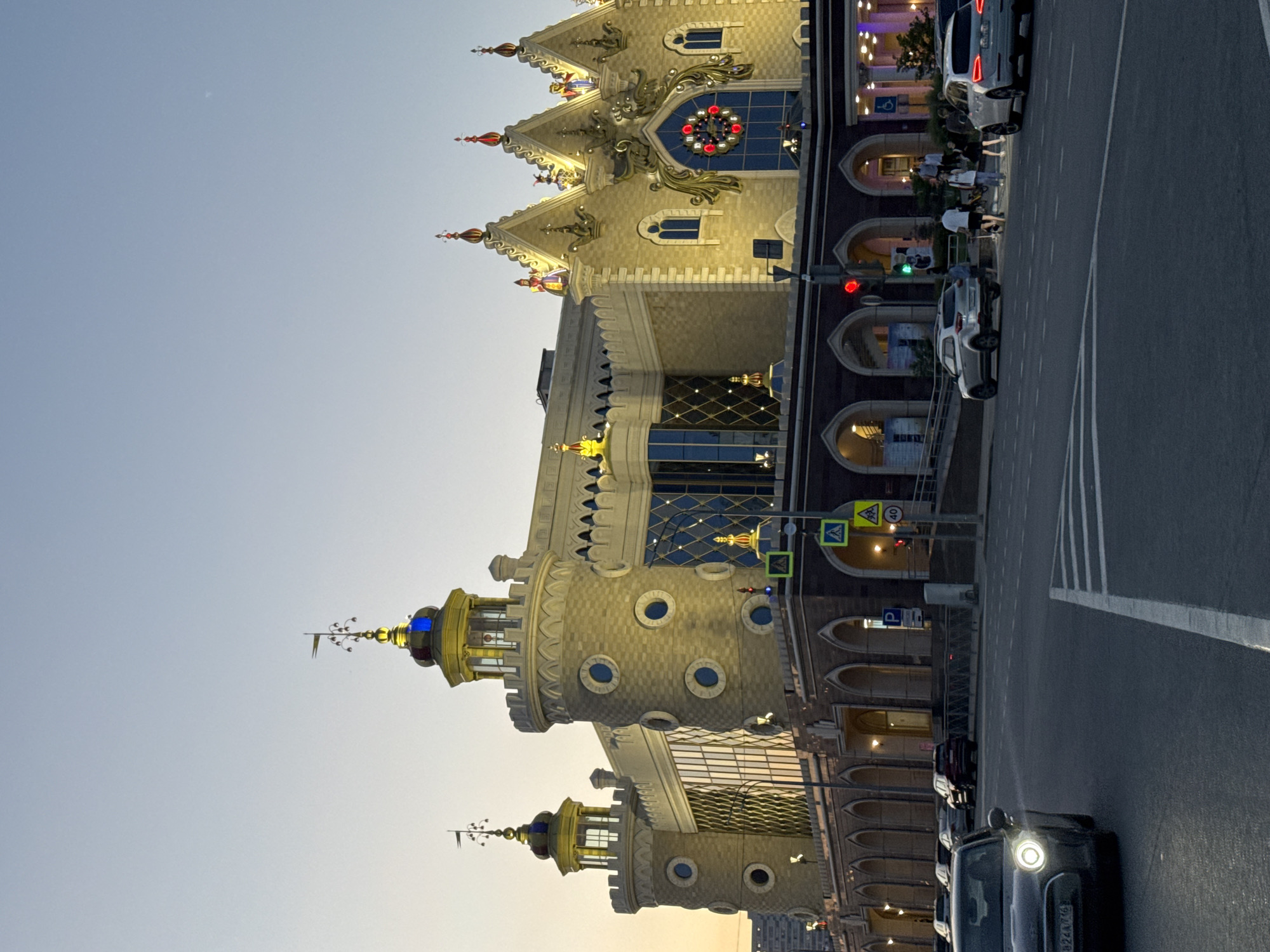 The image shows the Palace of Farmers in Kazan, Russia at dusk. The ornate building is prominently displayed on the left side of the image. A street runs along the right side of the building with parked cars. Pedestrians are crossing the street at a crosswalk. The sky is visible in the upper left portion of the image.