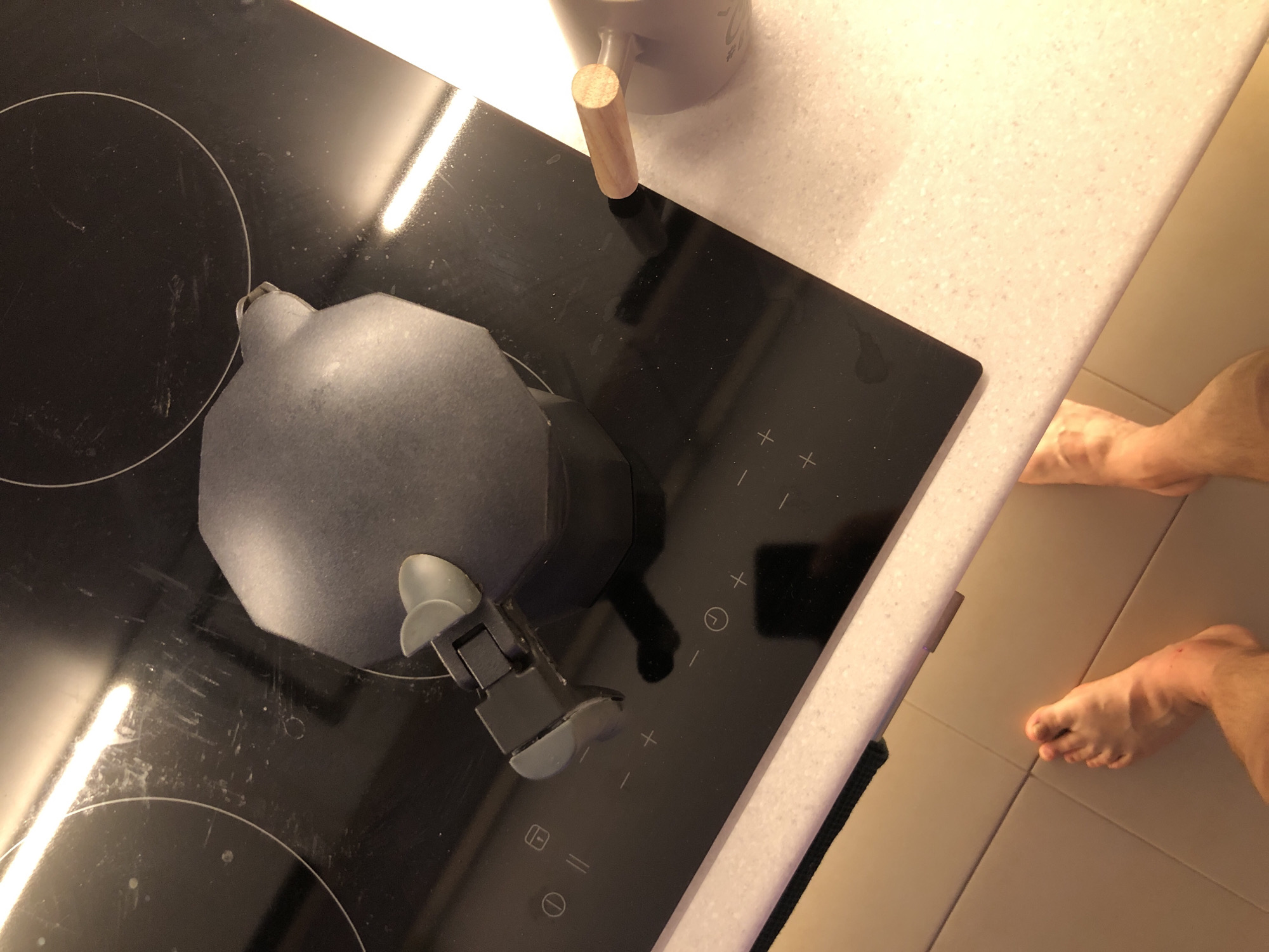 An overhead shot captures a domestic kitchen scene, likely in Saint Petersburg, Russia, featuring a black induction cooktop and surrounding surfaces. A dark, octagonal cooking pot, resembling a moka pot, is positioned on the front-left burner. The glass surface of the cooktop shows visible scratches and smudges, reflecting bright light sources. Control symbols for heat adjustment (+/-), timer, and power/boost functions are visible on the right side of the cooktop.
To the upper right of the cooktop, a light gray ceramic mug with a light-colored wooden handle rests on a speckled, light beige countertop. Faint, illegible text is visible on the side of the mug. On the far right of the frame, the bare feet of a person are visible on a light brown tiled floor, suggesting someone is standing in front of the counter, perhaps preparing a meal or coffee. The left foot is mostly visible, while only the toes and part of the arch of the right foot are seen. The scene depicts a quiet, everyday activity, possibly during morning or daytime.