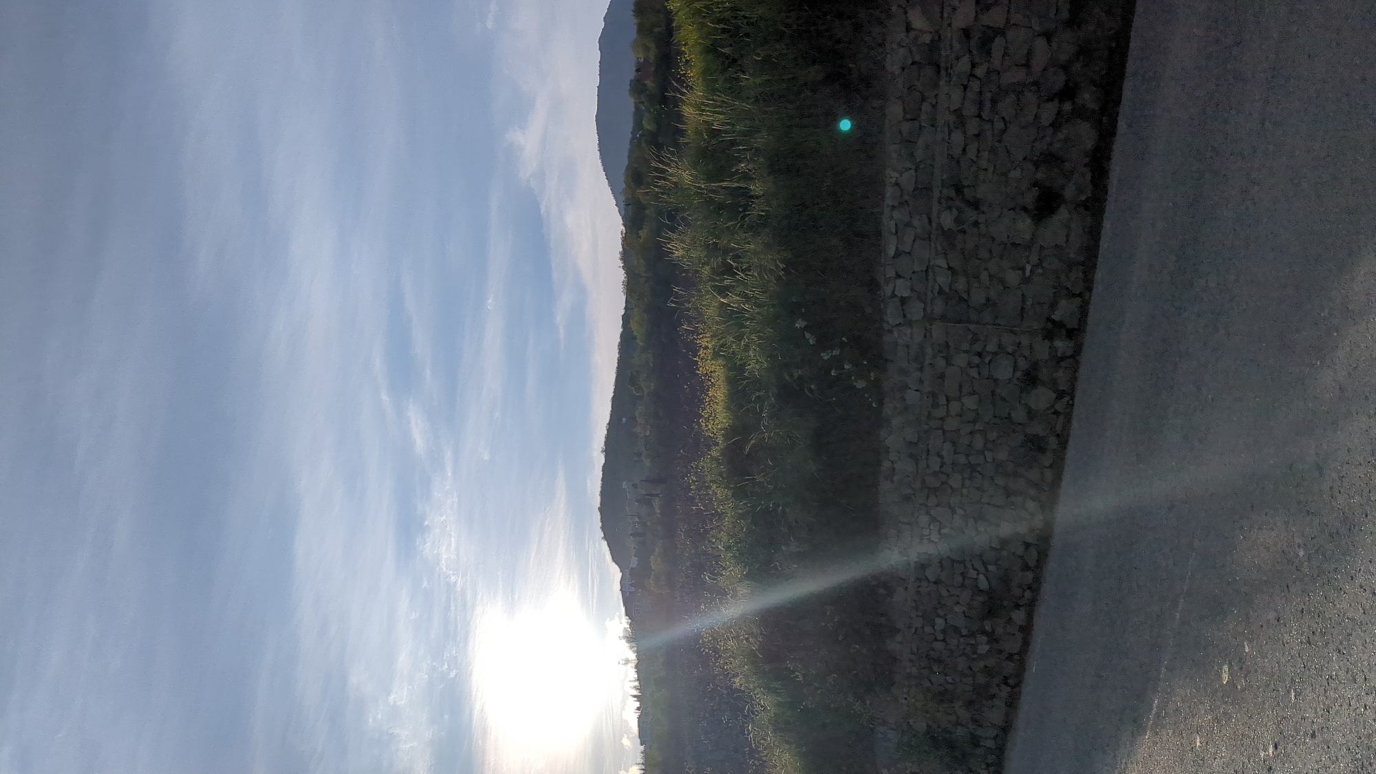 The image shows a landscape with a sky filled with wispy clouds. In the middle ground, there is a hillside with vegetation and a stone wall along the edge. To the right of the wall is a paved surface, likely a road or path. The sun is positioned low in the sky, casting a bright glow and some lens flare. The location is unspecified.