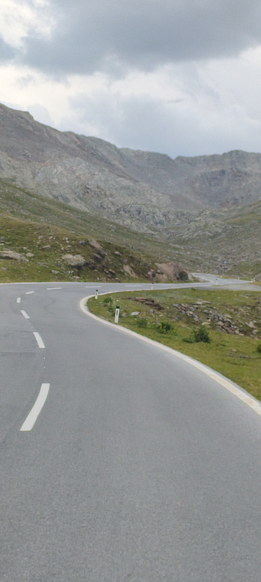 This image depicts a winding mountain road in the city of Sölden, Austria. The road is paved and has white dashed lines marking the lanes, as well as a continuous white line along the edge. On the right side of the road, there are white and orange marker posts placed intermittently. The surrounding environment is mountainous, with rocky slopes and patches of green grass and vegetation. The sky is overcast with gray clouds. There are no people or vehicles visible in the image.