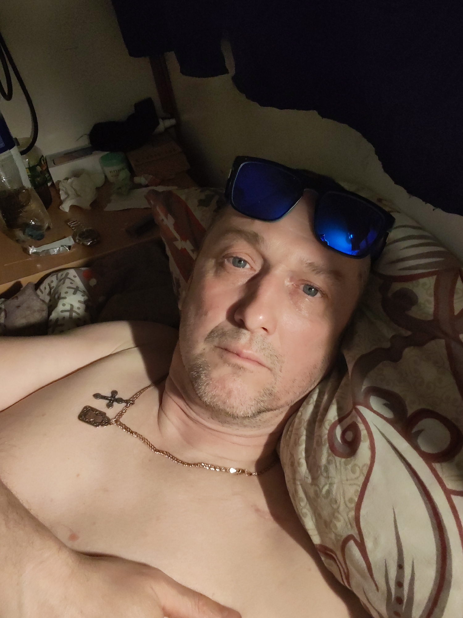 A man, appearing middle-aged with short, light-colored hair, light blue eyes, and light stubble, is depicted in a close-up, chest-up shot. He is lying down, shirtless, looking directly at the camera. He wears a gold-colored chain around his neck, featuring two pendants: a small dark cross and a rectangular, possibly metallic, charm. Blue-mirrored sunglasses are perched on his forehead, reflecting ambient light. A small, reddish mark is visible on his left chest.
He is positioned on a pillow with a light background and a distinctive dark red/brown patterned design. Another patterned fabric, possibly a blanket or sheet, is visible to his left. The overall lighting is dim, suggesting an indoor setting during the evening or night.
To the left of the man, in the background, is a cluttered surface. Visible items include a clear plastic bottle containing a brown liquid, another bottle with a green cap, crumpled white tissues or paper, and other indistinct objects. A dark cable or strap hangs vertically in the far upper left. Dark fabric, possibly clothing, is also visible in the background, suggesting a personal space such as a bedroom. There is no visible text in the image.