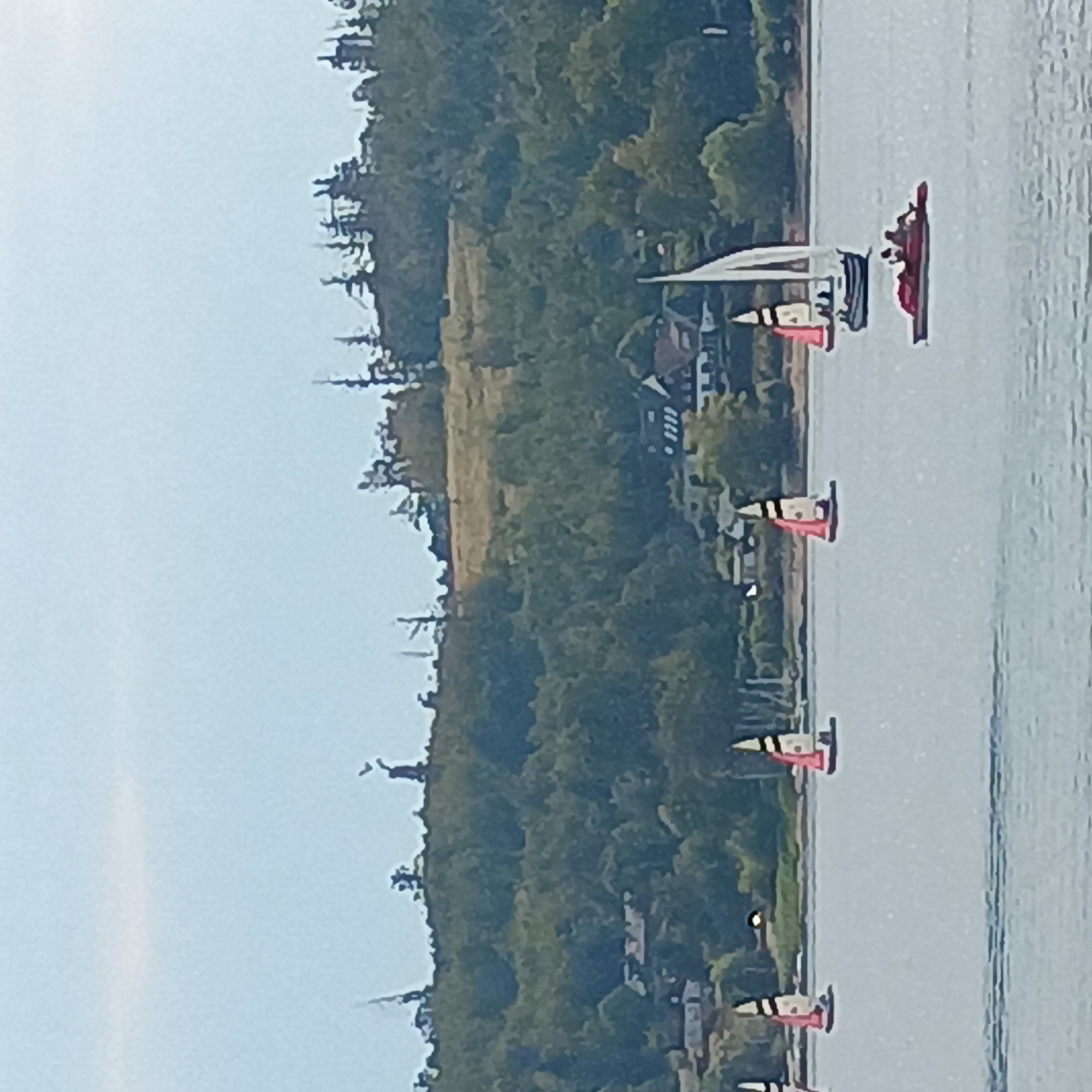 The image depicts a body of water, likely a lake, with several sailboats in the foreground and middle ground. The sailboats have colorful sails, with some featuring red and black striped patterns. A small red pedal boat with several people is also visible. In the background, a tree-covered hill with scattered buildings and a distant windmill-like structure dominates the landscape. The sky is light blue with some wispy clouds. The scene appears to be a recreational area, possibly in the vicinity of Möhnesee, Germany, known for its lake and surrounding natural beauty.