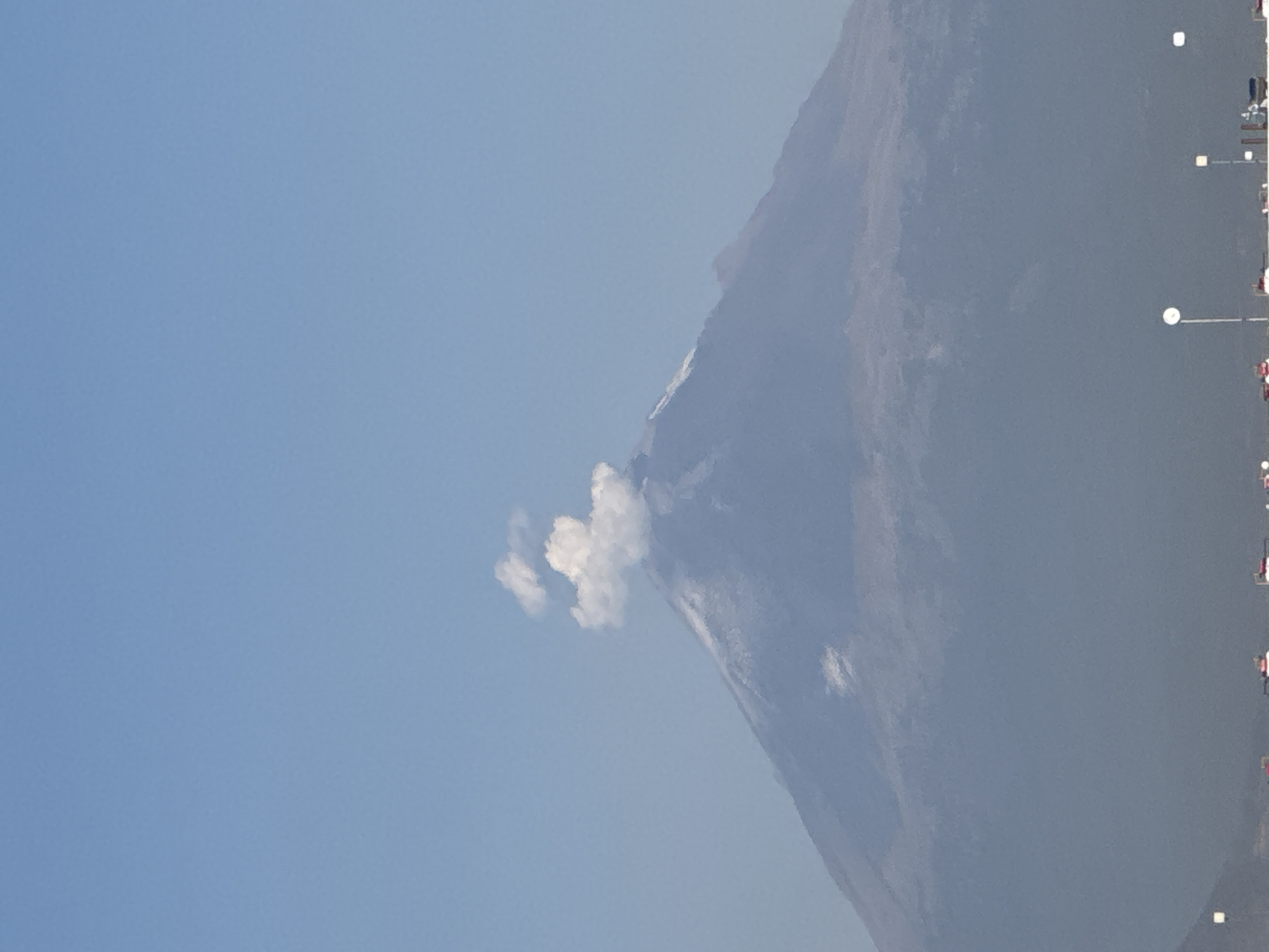 This vertically oriented image captures a majestic mountain or volcano, likely Popocatépetl, rising on the right side of the frame against a clear, bright blue sky that fills the left. A prominent plume of white smoke or steam emanates from the mountain's peak, indicating volcanic activity. The mountain's slopes are a muted greyish-blue, suggesting its vast scale and distance, with subtle textural variations. Along the lower right edge, a series of streetlights with glowing white tops are visible, alongside what appear to be distant structures or vehicles, suggesting a human settlement or road at the mountain's base. The scene is brightly illuminated, indicating it was taken during daylight hours with good weather conditions. No specific human activity is depicted, apart from the implied presence of infrastructure. There is no visible text in the image. The context provided places the view from San Francisco Ocotlán (Ocotlán), Mexico.