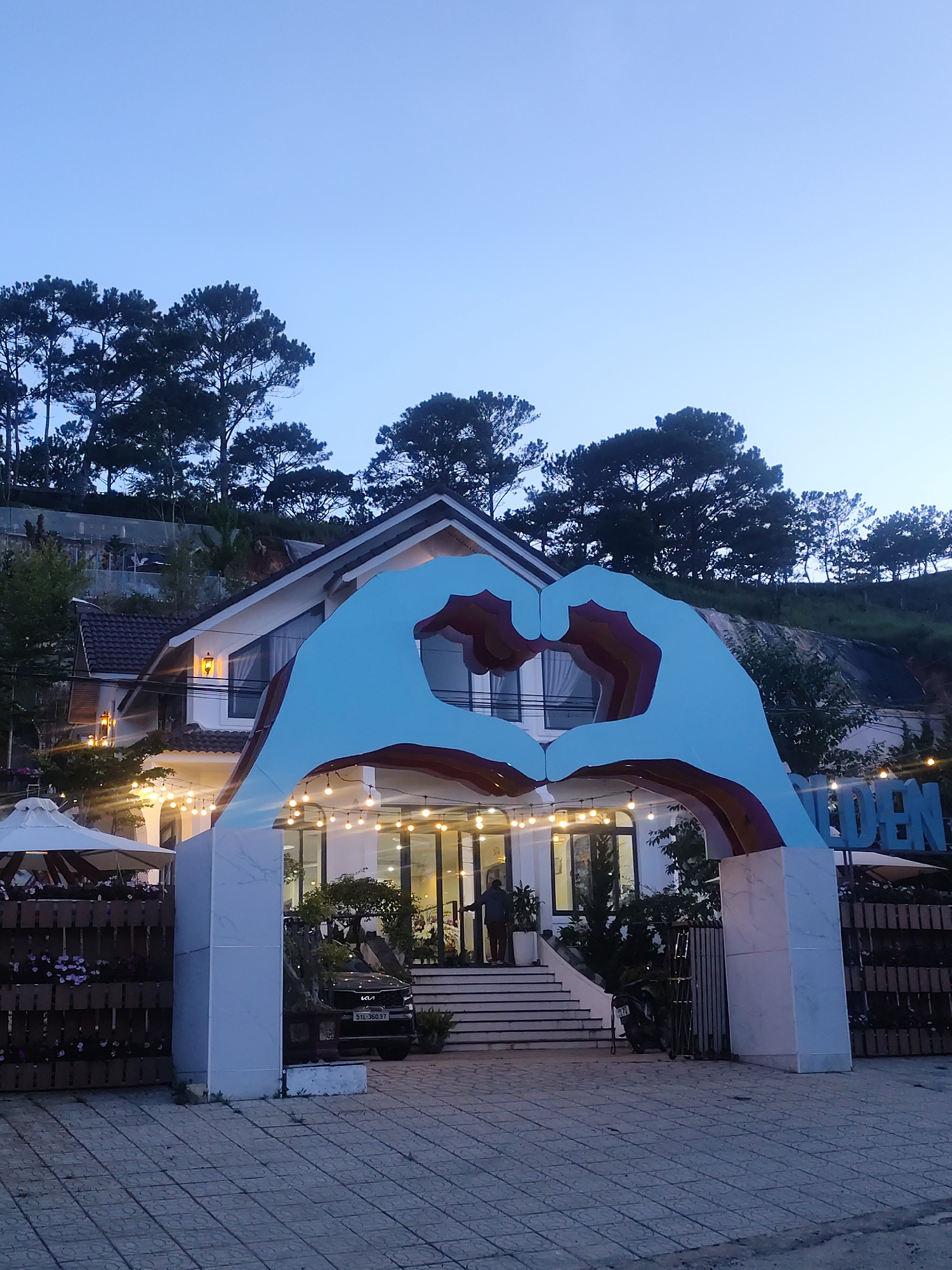 The image depicts a building with a large heart-shaped archway in front of it, at dusk. The archway is composed of two large hands forming a heart, with visible layers of blue, red, and brown on the inside curves. The building behind the archway is white with a dark roof and features large windows and string lights that are illuminated. There are steps leading up to the entrance of the building, and a person is standing at the top of the stairs. To the left of the entrance, there is a white umbrella and a wooden fence with planters. To the right, a dark-colored car with license plate "51L.380.37" is parked. A motorcycle is also visible near the entrance. The scene is set against a backdrop of lush green trees and a gently sloping hillside, characteristic of a mountainous or hilly area. The sky is a clear, pale blue, indicating the transition from day to night. The overall mood is serene and welcoming, possibly suggesting a resort or a place of accommodation. Visible text on a sign to the right of the archway reads "GOLDEN". The location is identified as Lạc Dương, Vietnam.