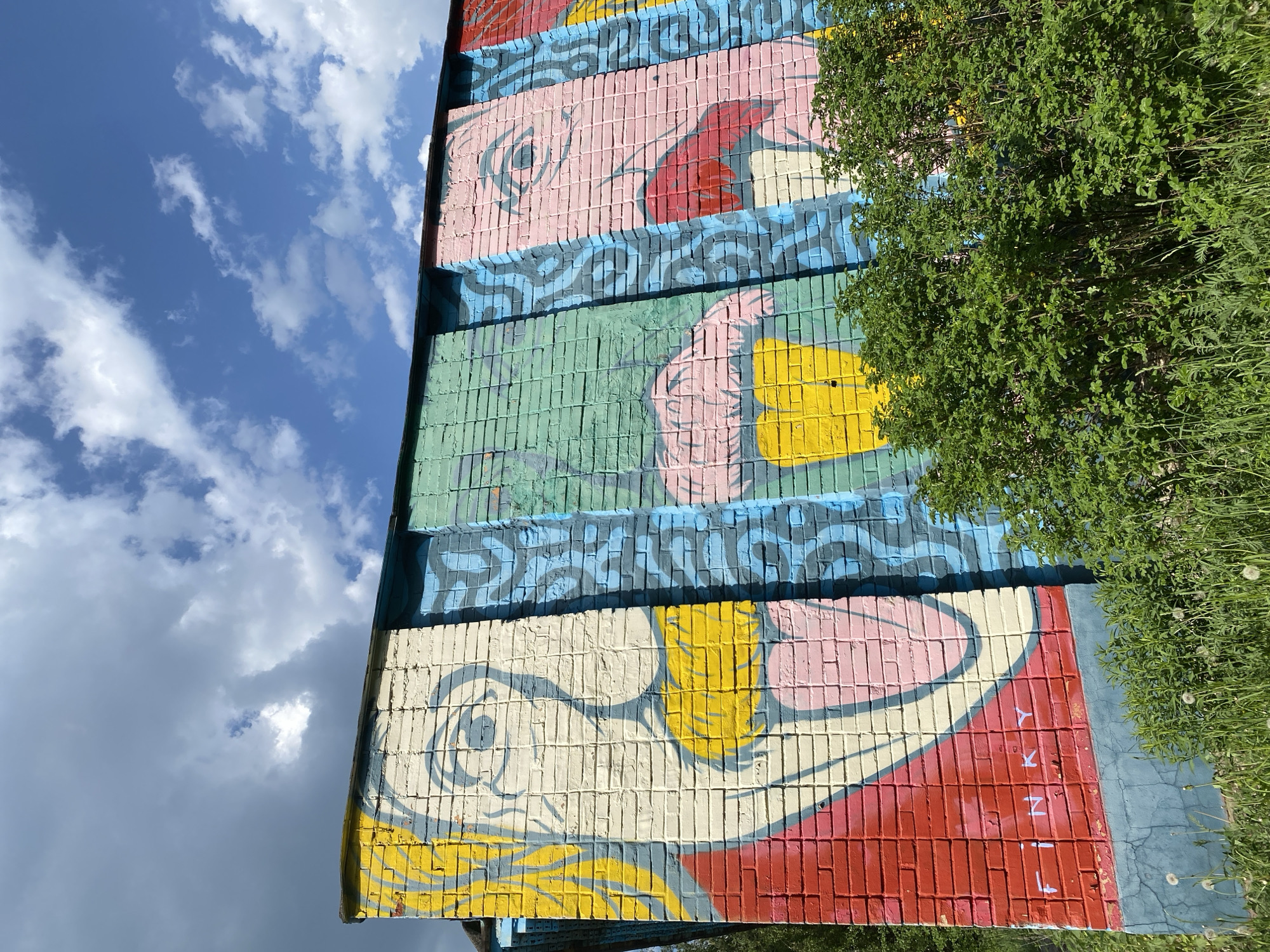 A low-angle shot captures a building's brick exterior adorned with a mural. The mural features a stylized representation of faces, with the bottommost face painted in shades of yellow, red, and white, and the letters "Lina" written vertically in red over a red and white background. The face above this is green with a pink hand. Above that is a face that is pink, red, and white. Blue abstract shapes frame each face, dividing the sections of the mural. The mural is partially obscured by a bush on the right. The sky, which appears to be overcast, is visible above and to the left of the building.
