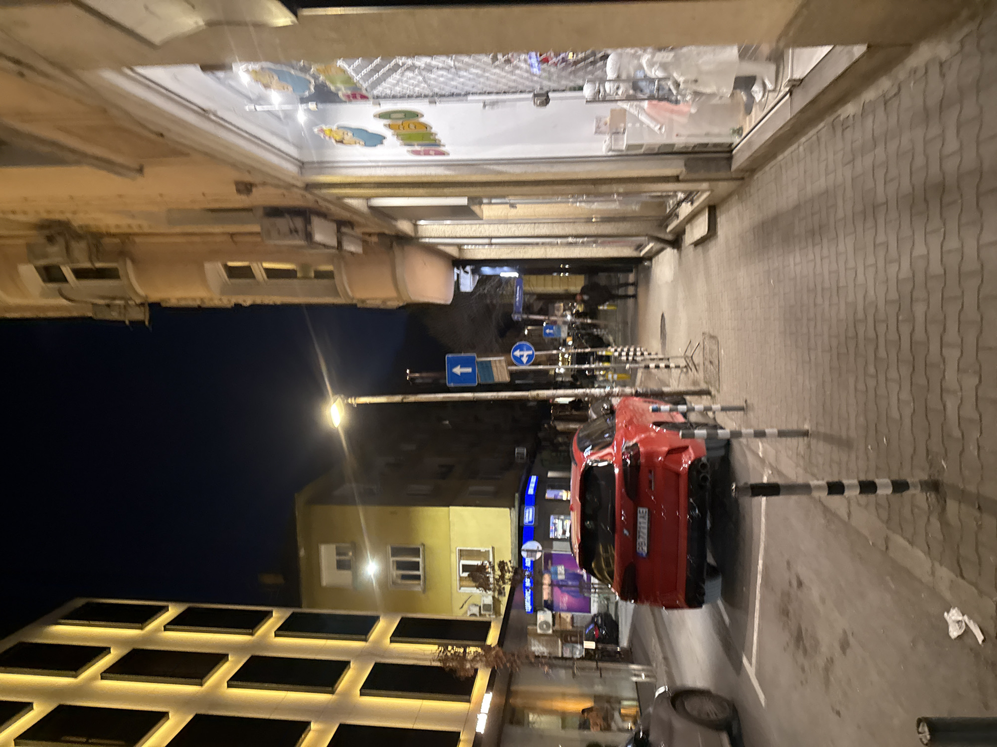 The image captures an urban street scene at night in Sofia, Bulgaria. The perspective is from a paved sidewalk, looking down a street illuminated by artificial light.
In the immediate foreground on the right, a grey interlocking brick sidewalk is visible, bordered by a series of black and white striped bollards that delineate the street. One bollard appears bent. A small piece of white paper refuse lies on the sidewalk near the bollards.
On the asphalt street, a red SUV, identified as a BMW X5 M series or similar performance model, is parked or stopped on the right side. Its rear faces the viewer, displaying Bulgarian license plate PB 7711 AE and quad exhaust pipes. Faint white parking lines are visible on the street surface. To the left of the red SUV, the front portion of a silver/grey sedan is partially visible, positioned further down the street.
A tall black street lamp pole stands near the red SUV, equipped with a bright white luminaire. Mounted on the pole are two blue circular road signs: one indicating "straight ahead" with an upward-pointing white arrow, and another indicating "turn left" with a white left-pointing arrow.
On the right side of the street, a multi-story building with a light beige facade features ground-level retail spaces. A large storefront window on the far right displays several white mannequins dressed in children's clothing, accompanied by colorful decorative elements including cartoon-like figures and the partially visible word "BABY."
On the left side of the street, a modern building with a distinctive facade composed of large, dark rectangular panels outlined by bright yellow LED strip lighting is prominent. Adjacent to it stands a multi-story yellow building with multiple illuminated windows, indicative of residential use. Below the yellow building, a storefront with blue illuminated signage clearly displays "ЦВЕТАРСКИ МАГАЗИН" (Flower Shop) in Cyrillic.
In the mid-distance on the right sidewalk, several indistinct human figures are visible walking, appearing as dark silhouettes against the background lighting. The overall environment is a commercially active city street under a dark night sky.