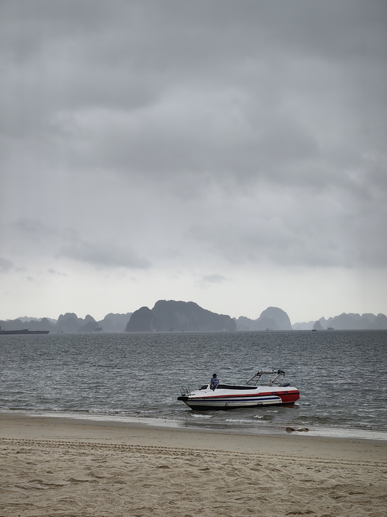 The image shows a beach scene in Bai Chay Ward, Vietnam, with a motorboat partially beached on the sand. The boat is white with red and blue stripes and a man sits on its edge. In the background, the sea stretches towards a horizon dotted with limestone karsts shrouded in mist. The sky above is overcast with grey clouds. The sand on the beach shows tire tracks. The sea is grey and choppy.