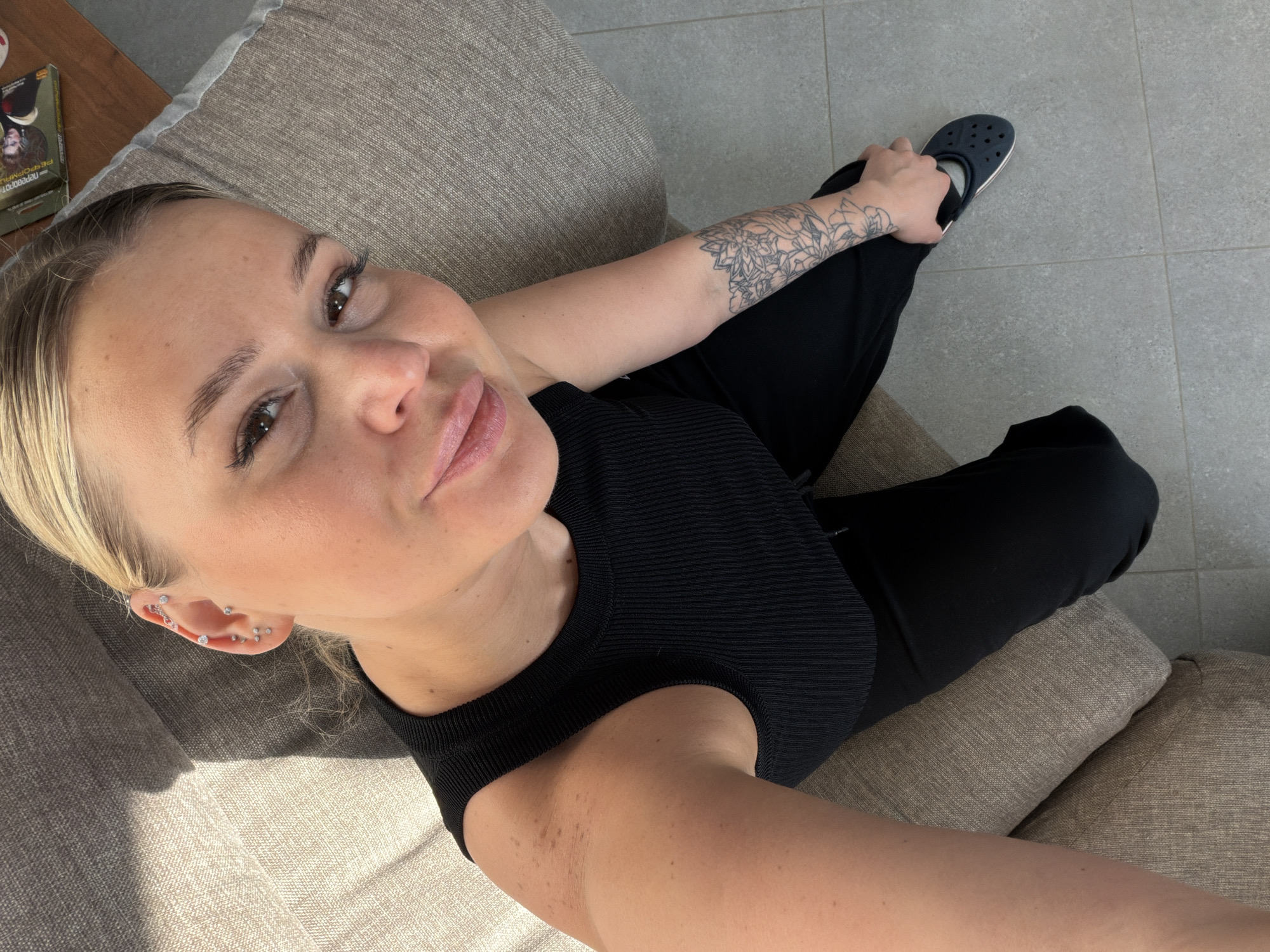 The photograph shows a blonde-haired woman in a black top and black pants sitting on a beige couch. She is positioned at an elevated angle, looking up toward the camera, with a tattoo on her left arm. She is wearing multiple earrings in her left ear. A navy blue clog is located near her foot. The background includes a neutral-colored tiled floor and a wooden surface with a book. The location is not identifiable as Sarapul, Russia from the image.