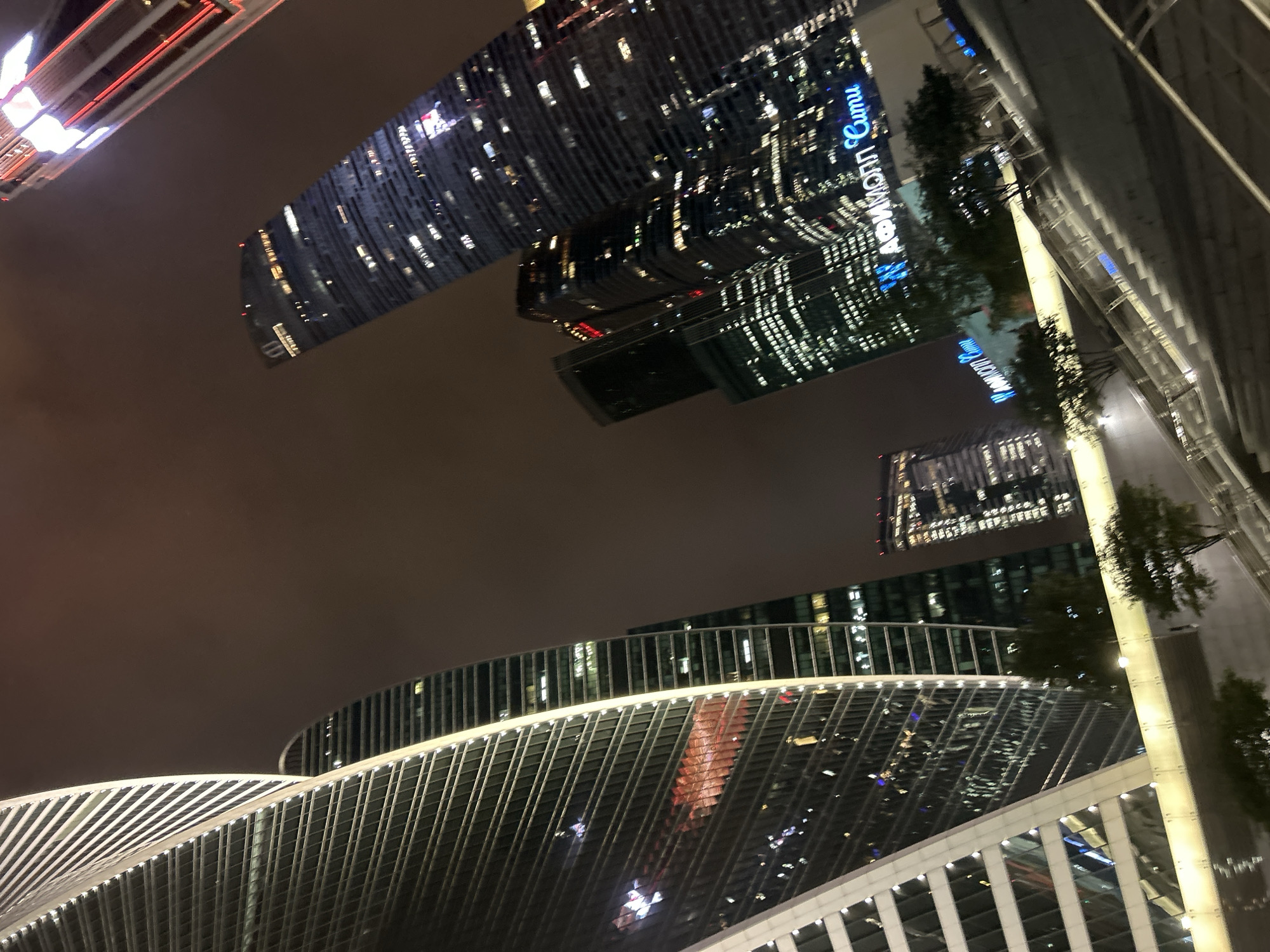 An exterior, eye-level, wide shot captures a night view of several modern skyscrapers in an urban setting. The sky is dark and cloudy, suggesting nighttime or poor weather. The buildings are illuminated with various lights, some with architectural lighting highlighting their structures and others with illuminated windows indicating occupancy. The prominent building on the left is a twisted skyscraper with a reflective glass facade. Other buildings in the background range in height and architectural style, with visible illuminated signs. In the foreground, there is a staircase leading upwards, and some trees are visible at the base of the buildings. The overall impression is one of a modern, bustling city at night.