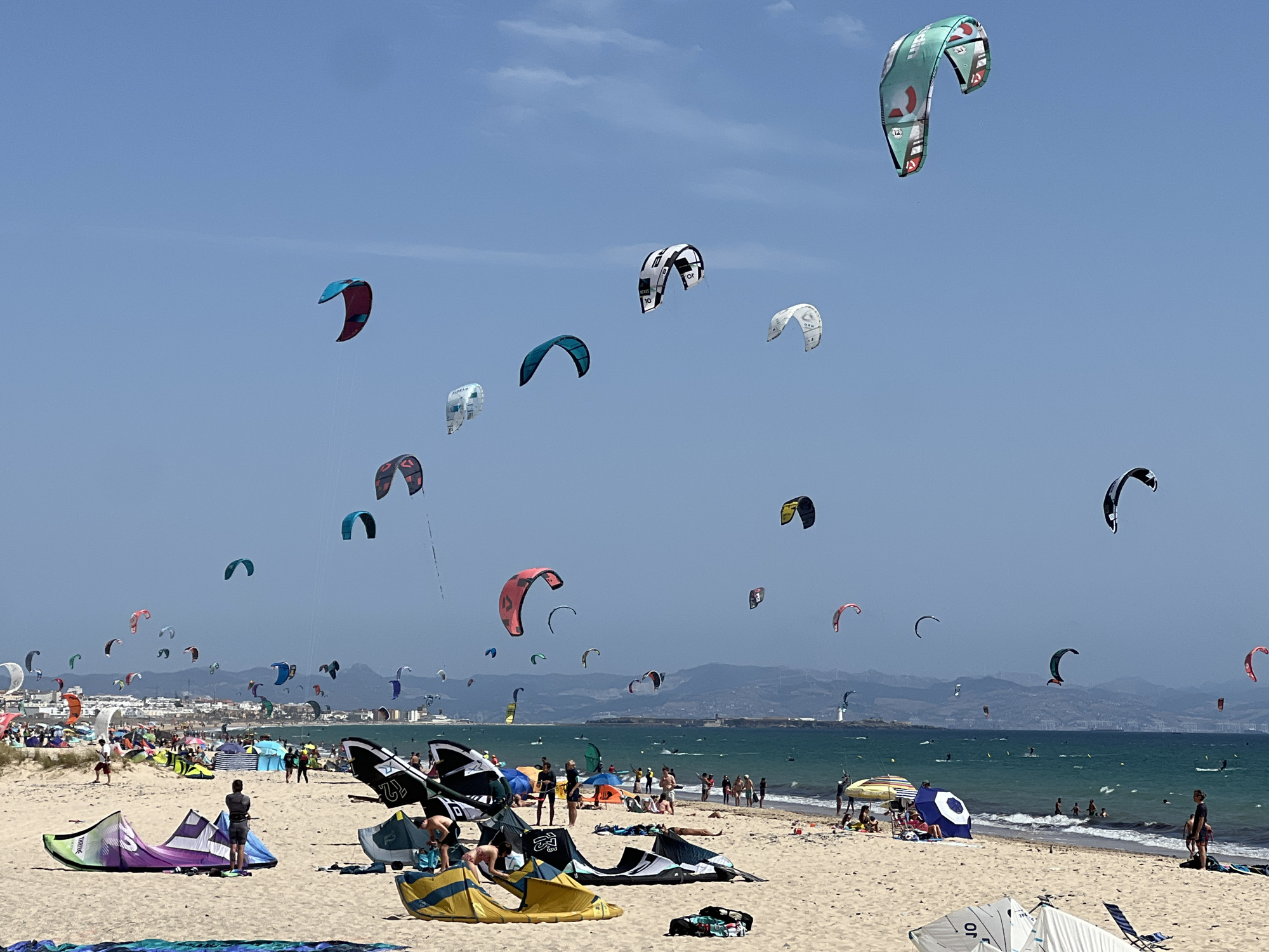 This is an aerial photograph, taken from a high vantage point, looking down on a crowded beach in Tarifa, Spain, on a bright, sunny day. The sky is a clear blue, with a few wispy clouds. The sea is a vibrant turquoise, with small waves breaking on the shore.
The beach is packed with people, most of whom are involved in kitesurfing. There are dozens of kites in the air, in a variety of colors and sizes, with their strings visible against the sky. Some kites are very close to the water, while others are higher up. Several kites are in various stages of being launched or landed on the sand.
On the beach, there are many colorful tents and umbrellas, providing shade for the sunbathers. People are seen walking, sitting, and playing in the sand. In the water, there are a few swimmers and kitesurfers.
In the background, there is a town with white buildings and a lighthouse. The town appears to be nestled against a backdrop of rolling hills. The overall impression is one of a lively and active beach scene, with kitesurfing being the main activity.