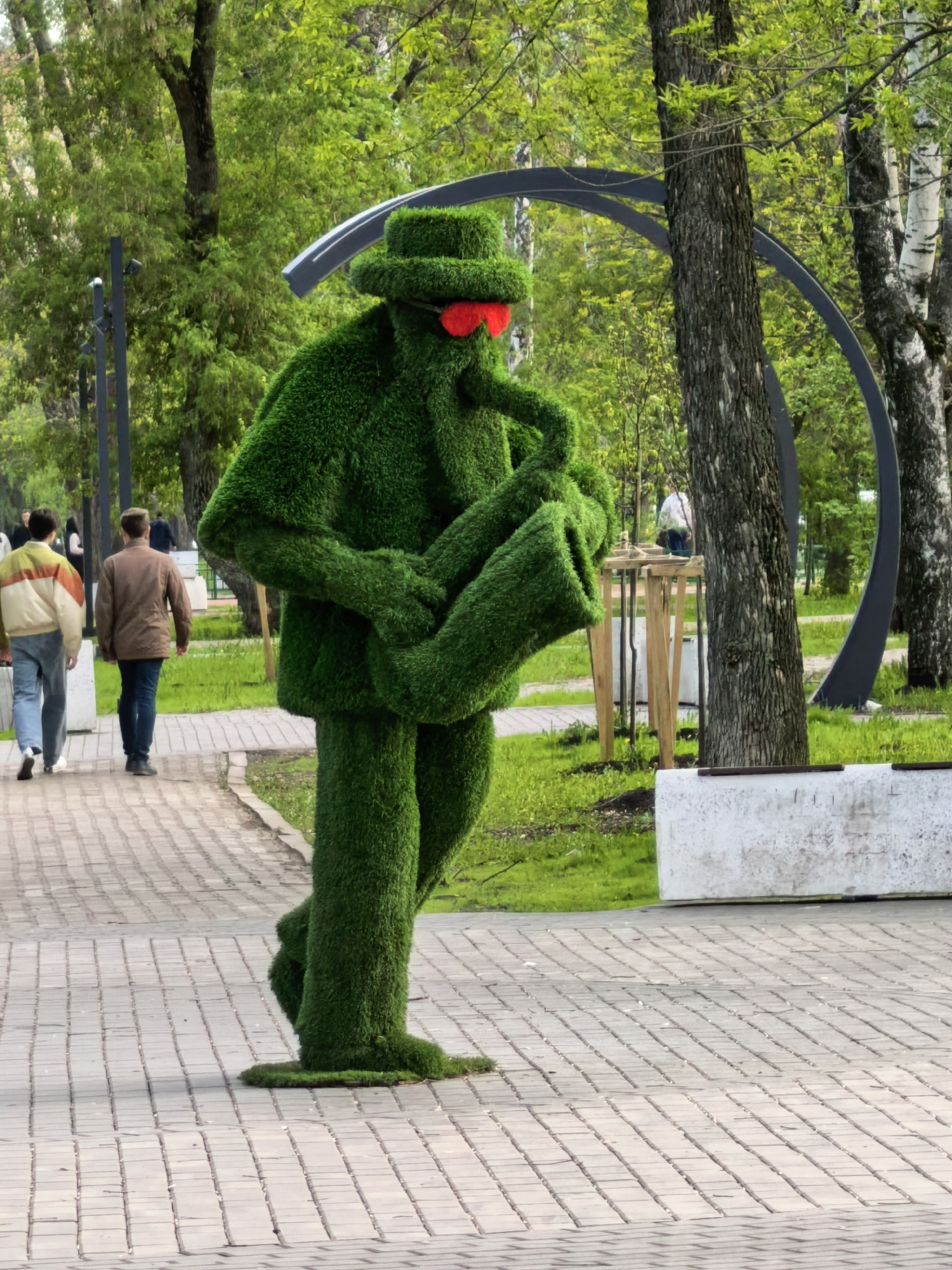 In Sterlitamak, Russia, a whimsical outdoor sculpture of a person completely covered in artificial green grass stands on a brick-paved pathway. The grass-covered figure sports a hat and red sunglasses and holds a saxophone, also made of the same green grass material. Two pedestrians stroll along the pathway in the background to the left of the sculpture, while various trees and a curved metal architectural element add depth to the backdrop. A section of grass and a white concrete structure are visible to the right of the sculpture.