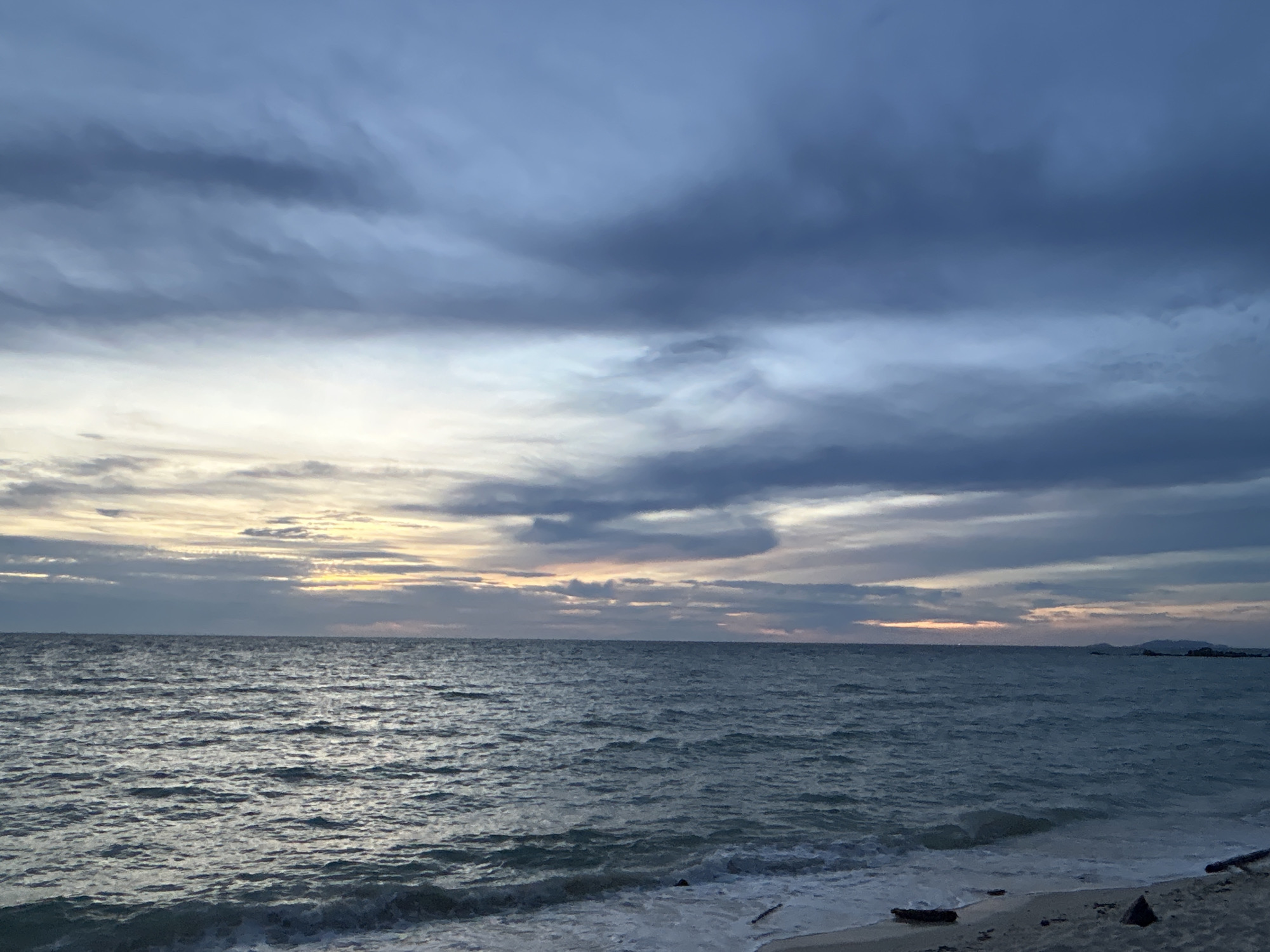 The image depicts a serene beach scene at dusk on Koh Phangan, Thailand. The sky is filled with dramatic, heavy clouds in shades of dark blue and grey, with a subtle band of warm orange and yellow light peeking through near the horizon, indicating the setting sun. The ocean is choppy, with small waves breaking on the sandy shore. In the foreground, the sand is littered with a few pieces of driftwood. In the distance, faint outlines of landmasses or islands can be seen against the twilight sky. There are no people or animals visible in the frame. The overall atmosphere is tranquil and slightly moody due to the cloudy sky.