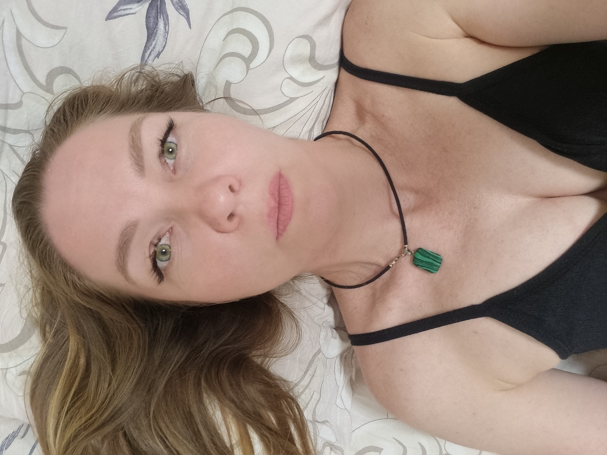 A woman is lying on her back, facing the camera. She has light skin, green eyes, and light brown hair that is partially visible in the lower left corner. She is wearing a black spaghetti strap top and a black choker necklace with a green pendant. The background is a white patterned bedsheet. The location and details surrounding the city of Chaikovsky, Russia, are not discernible from the visual data provided.