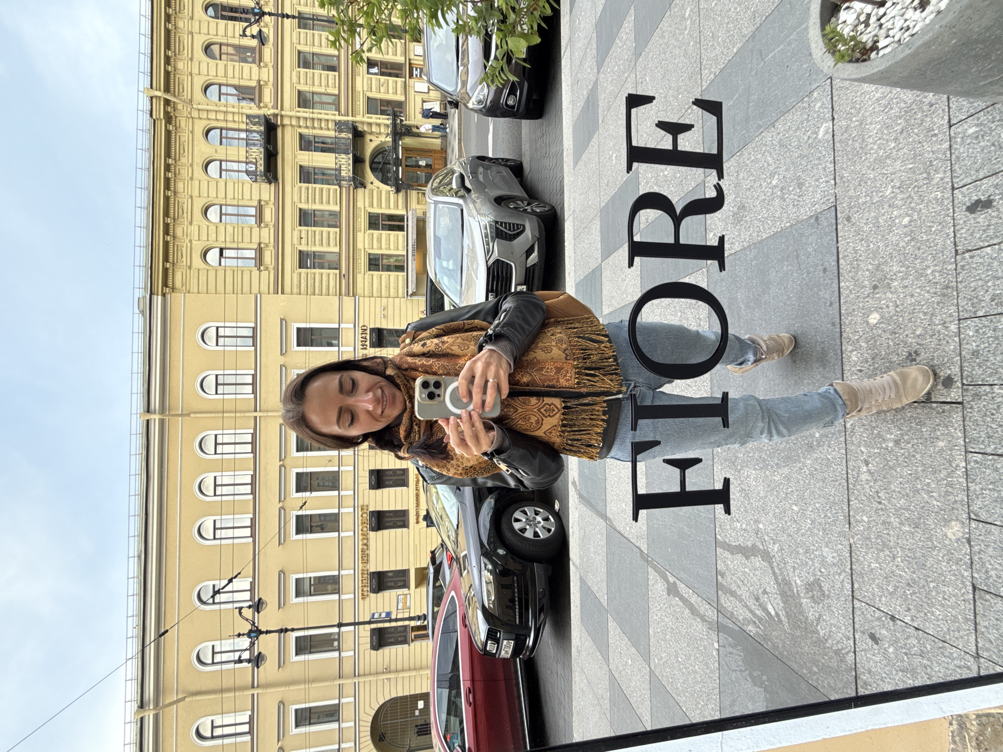A woman is taking a selfie with her phone in front of a yellow building. She is wearing a patterned scarf, a dark jacket, and blue jeans. She has long brown hair and is smiling. Large black letters spelling "FIORE" are visible on the pavement. Several cars are parked on the street behind her, and more buildings are visible in the background. The setting appears to be a city street with architectural details that suggest a European location, consistent with Saint Petersburg, Russia. The lighting suggests it is daytime, possibly overcast. The woman's expression and the selfie suggest a casual and possibly tourist activity.
