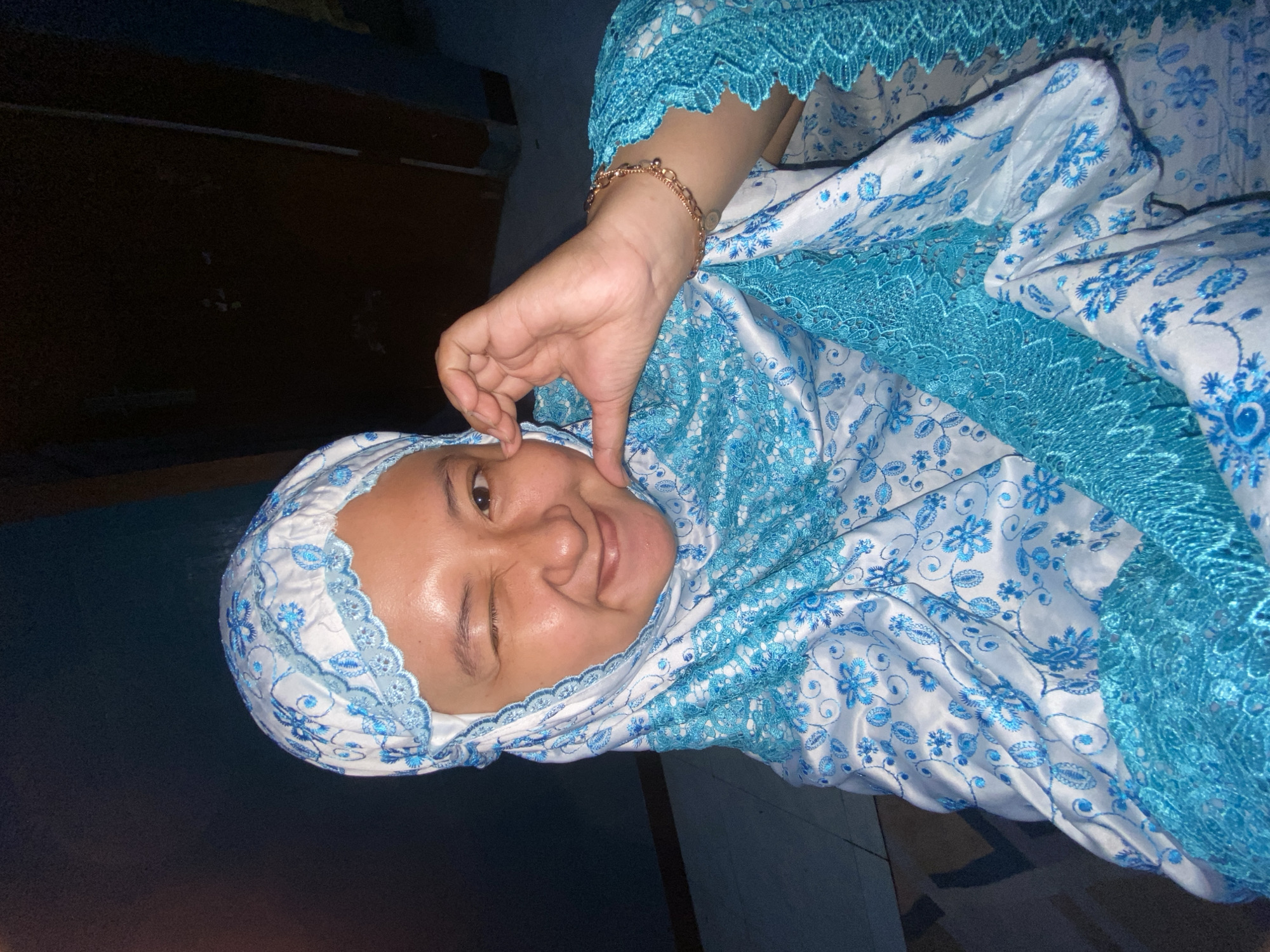 A young Indonesian woman is pictured indoors, possibly in her home in Gelam, Indonesia. She is wearing a white hijab adorned with blue floral embroidery and a matching blue lace trim. The woman is winking and making a heart shape with her hand, touching her cheek. She has a bracelet on her right wrist. The background is dark, suggesting it may be evening or nighttime, with a hint of a doorway visible. The floor appears to be tiled.