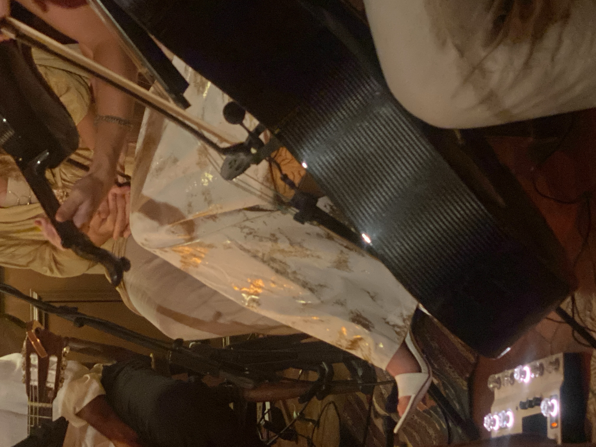 A canted, close-up shot focuses on a performer playing an electric cello, wearing patterned pants and white high heels. A microphone and guitar are partially visible in the lower left corner. A white effects processor is in the lower right. The location is likely a performance venue in ตำบลเกาะพะงัน, Thailand.
