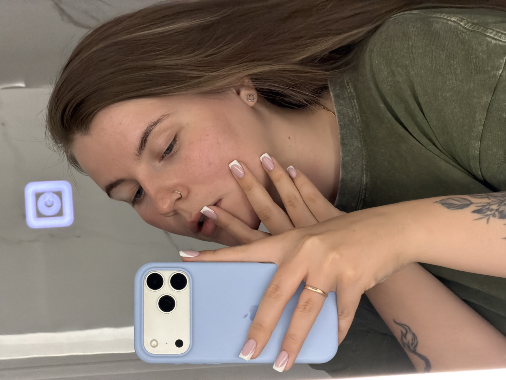 A person with long brown hair, wearing a green shirt and a gold ring, is taking a selfie in front of a mirror with a light blue smartphone case. The person has a nose ring and tattoos on their arm. The location is unknown; the request to include Chaikovsky, Russia, is not applicable based on the image content.