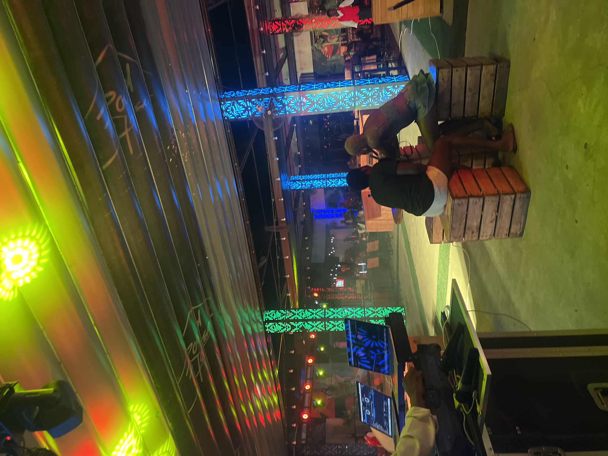 In Howayi, Nigeria, a lively outdoor night scene unfolds under a canopy illuminated by vibrant, multi-colored lights. Red, green, and yellow lights cast dynamic patterns on the corrugated metal ceiling and down onto the floor, which appears to be concrete covered with a thin layer of lime-green artificial turf.
The focal point in the foreground is a DJ setup, with two laptops and microphones positioned on a rack. A person, partially visible and wearing a light-colored sleeve, is operating the equipment. To the right, two individuals are seated on wooden crates. One, wearing a black t-shirt and white shorts, faces away from the camera. The other, with blonde hair, faces the first individual, their back to the viewer. They appear to be in conversation.
Further into the background, the venue is populated with more people, some seated at tables and others standing, suggesting a social gathering or event. Two tall, decorative columns, one lit with blue and the other with green, flank the central area, adding to the festive atmosphere. The overall environment suggests a bar, club, or entertainment venue.