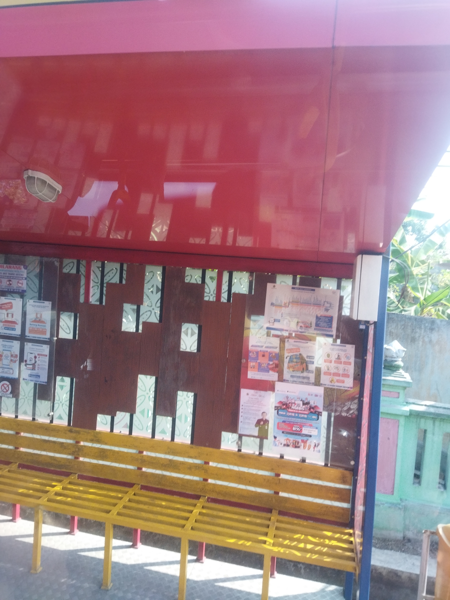 The image shows a red bus stop shelter with a yellow bench inside. Several posters are displayed on the back wall of the shelter. The bus stop is located in Begajah, Indonesia. The shelter has a red roof and red sides, with a blue trim. The bench is made of yellow metal bars. The posters are of various sizes and contain text and images. One poster has a graphic of a bus and the words "BIJIBRAJE". Another poster has pictures of people and the text "BUKAN MAIN....". The environment around the bus stop appears to be outdoors, with some greenery visible in the background. The ground is paved.