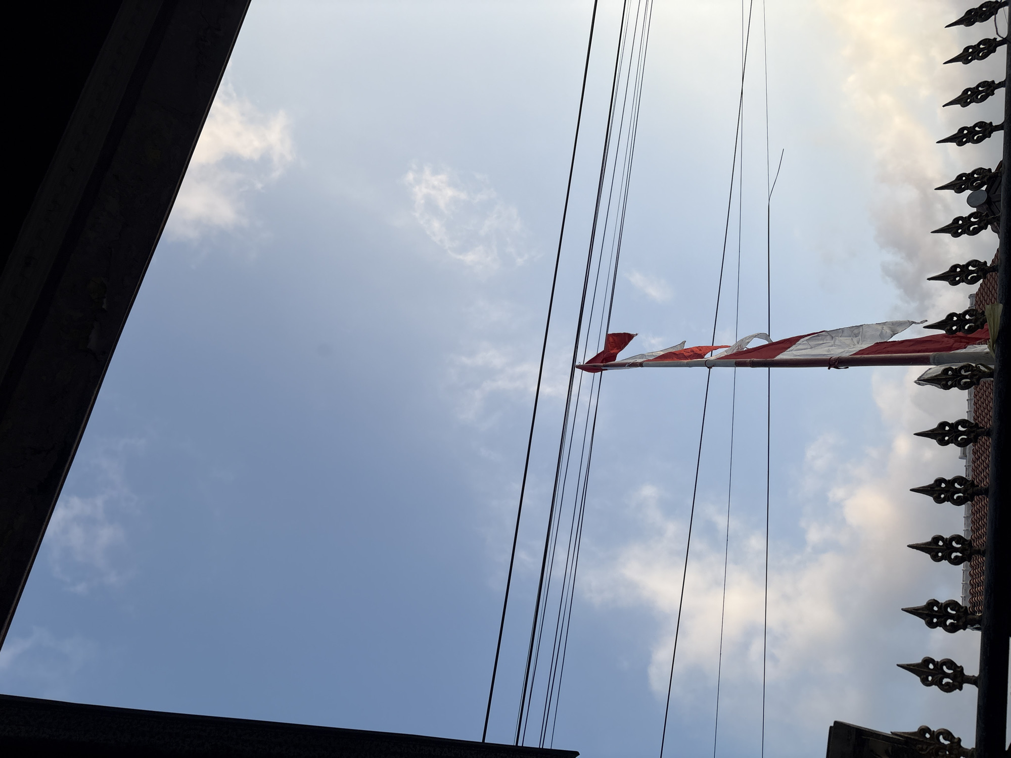 The image captures a view from under a dark overhang, looking up at a bright blue sky with scattered white clouds. In the mid-ground, a flagpole with an Indonesian flag, red and white, is positioned off-center to the right. Several dark power lines are strung horizontally across the sky in front of the flag and clouds. In the foreground, a black decorative metal fence with pointed finials is visible, bordering a tiled roof. The scene appears to be outdoors, possibly in a residential area in Palimanan, Indonesia, given the presence of the Indonesian flag and common architectural elements. There are no people visible in the image.