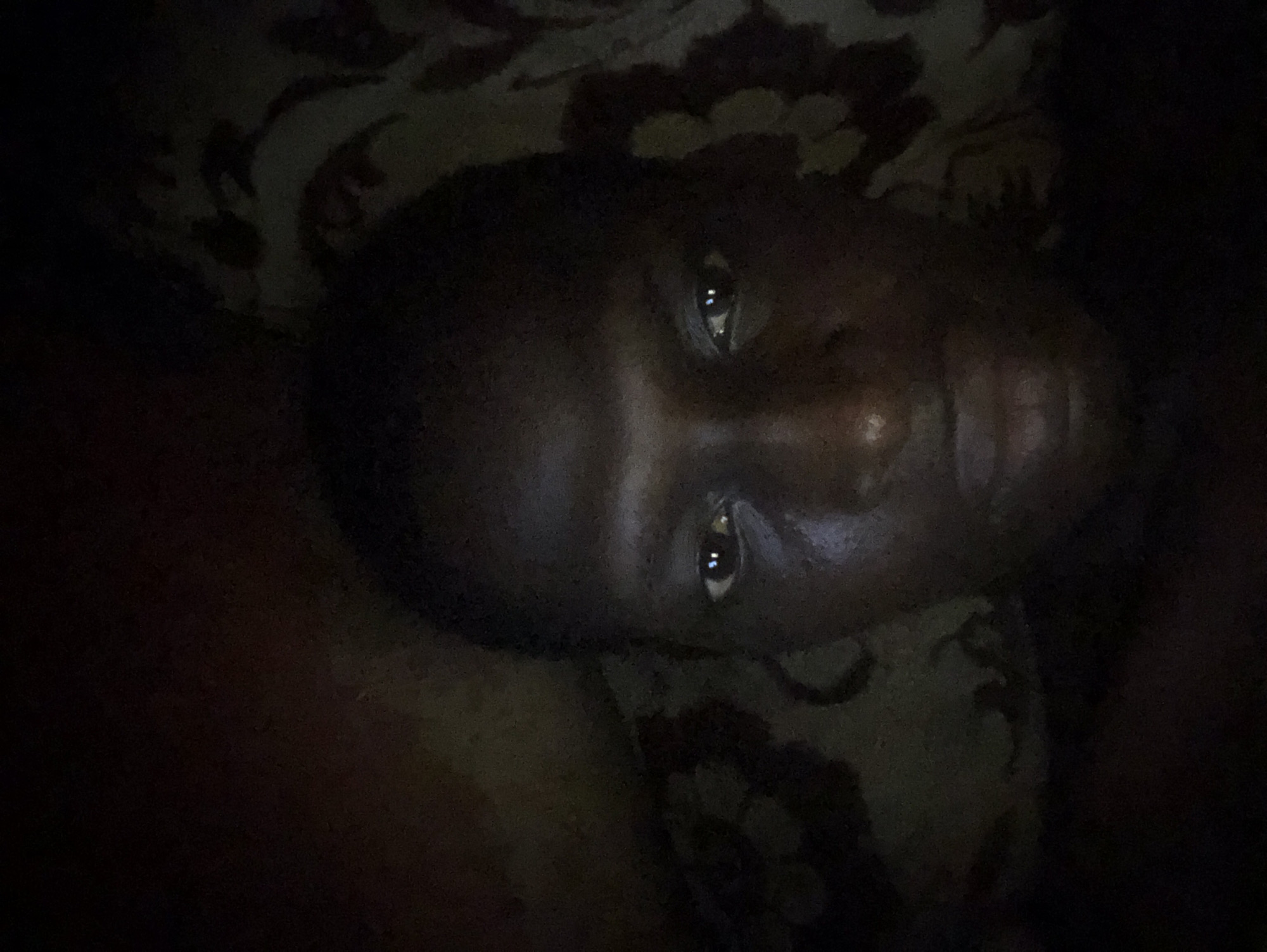 The image shows a person's face against a dark background with a patterned texture. The person has dark skin and their eyes are open, reflecting a light source. The image is poorly lit, making it difficult to discern specific details.