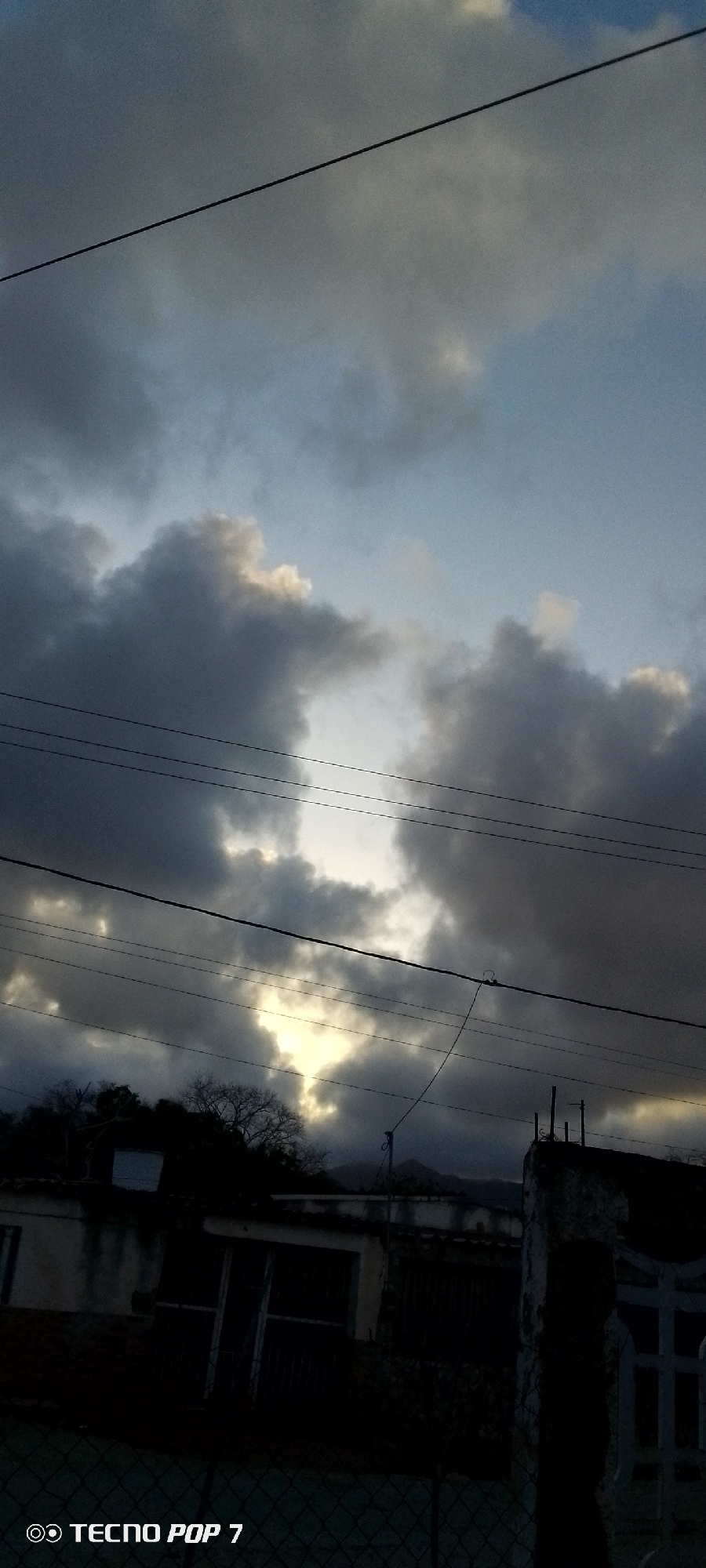The image depicts a cloudy sky with electrical wires in the foreground. Light is shining through a gap in the clouds. Below the horizon, there is a silhouette of a building with a tree. The photograph was likely taken in Altagracia, Venezuela, based on the prompt. The building appears to be dark, and its structure is obscured by shadow. A fence or wall is visible to the right. The text "Tecno Pop 7" is in the lower left corner of the image.