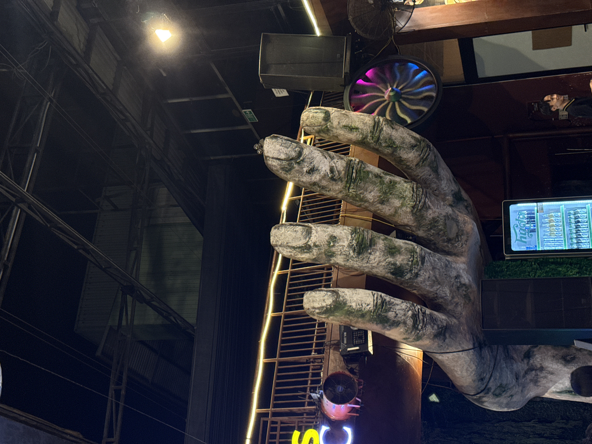 The image shows a large, moss-covered sculpture of a hand reaching upwards in an indoor setting. The hand is the central focus of the foreground. Behind the hand, a metal railing with yellow lights is visible, suggesting a walkway or balcony. To the left of the railing, a red fan is mounted, and neon letters that appear to spell "SC" are also visible. In the mid-ground, there is a dark speaker on the right and a colorful, spinning fan or light fixture above it. The background consists of the dark ceiling structure of the building, with exposed beams and lighting. On the far right, a man in a black shirt is standing, smiling. A brightly lit cooler with bottles of drinks is positioned on the lower left. The overall environment appears to be a dimly lit entertainment venue or bar. This scene is consistent with the entrance area of a popular establishment in Đà Nẵng, Vietnam.